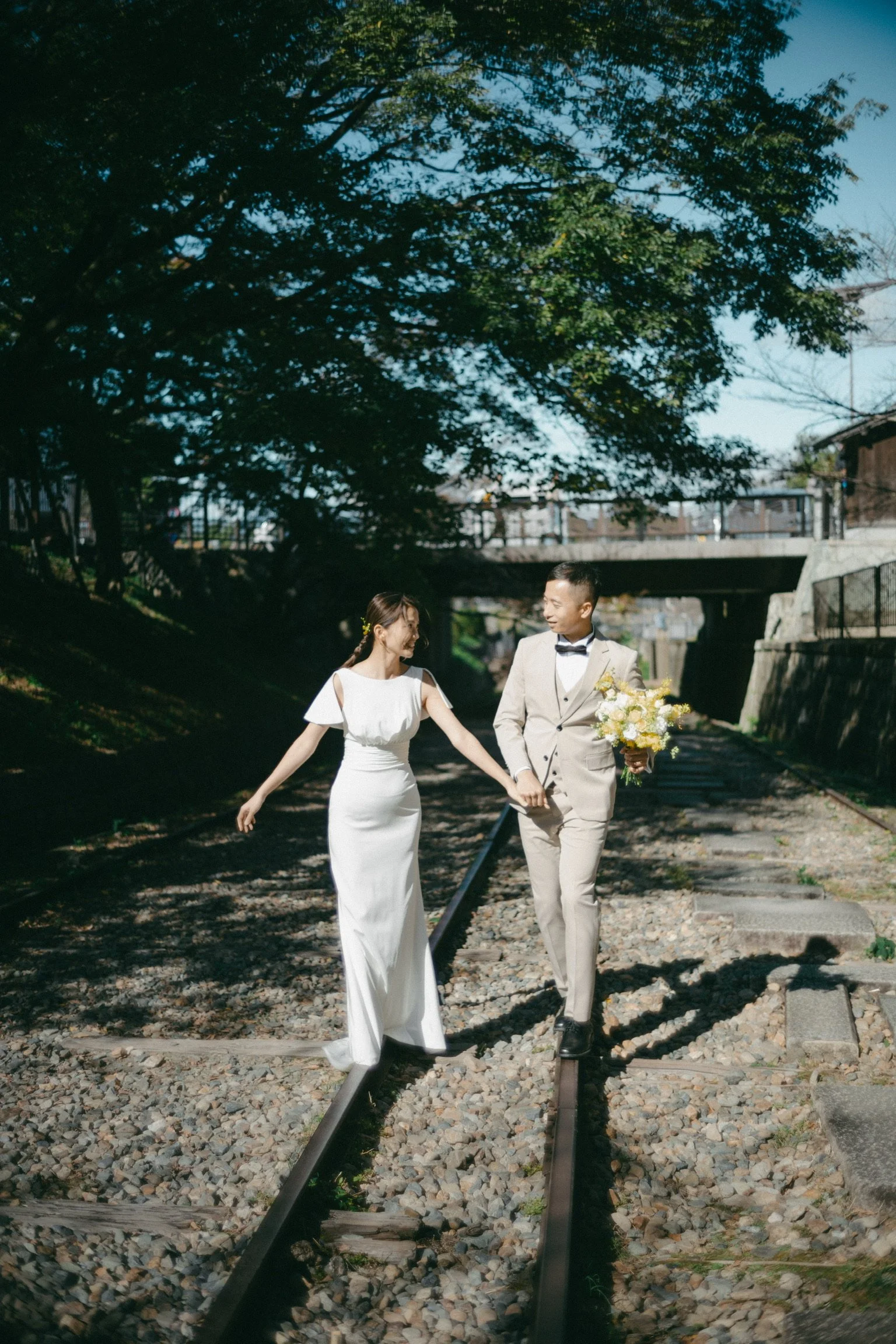 A bride and groom walking hand in hand on train tracks outdoors, with the bride in a white gown and the groom in a beige suit holding a bouquet. Large trees are in the background under a clear sky.