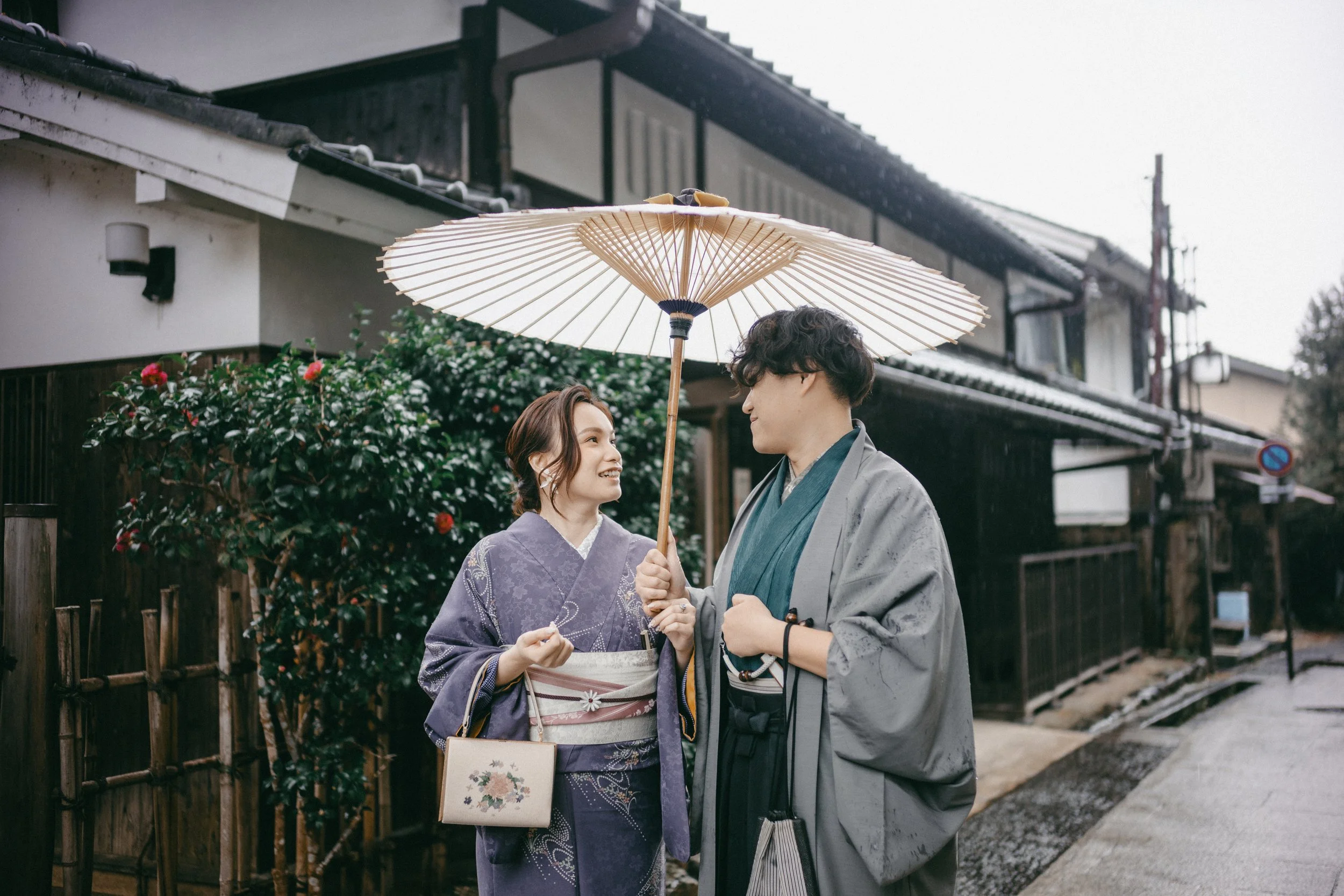 A young couple wearing traditional Japanese kimonos sharing a moment under a paper parasol on a rainy street.