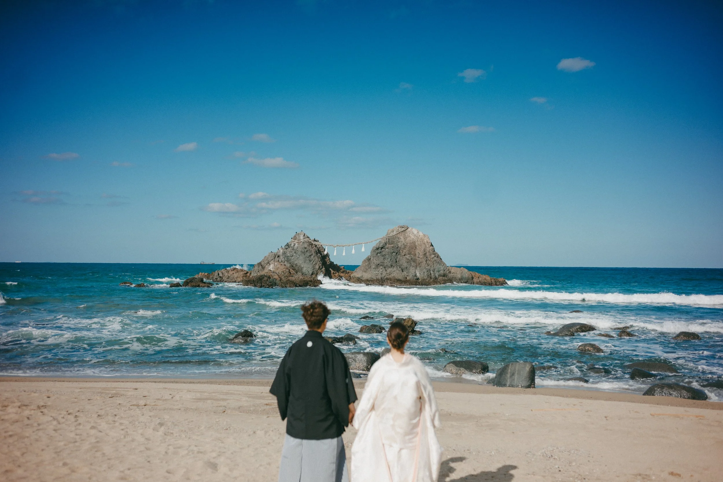 A couple dressed in traditional Japanese wedding attire hold hands and walk towards the ocean on a sandy beach, with rocks and a small shrine on the rocks in the background.