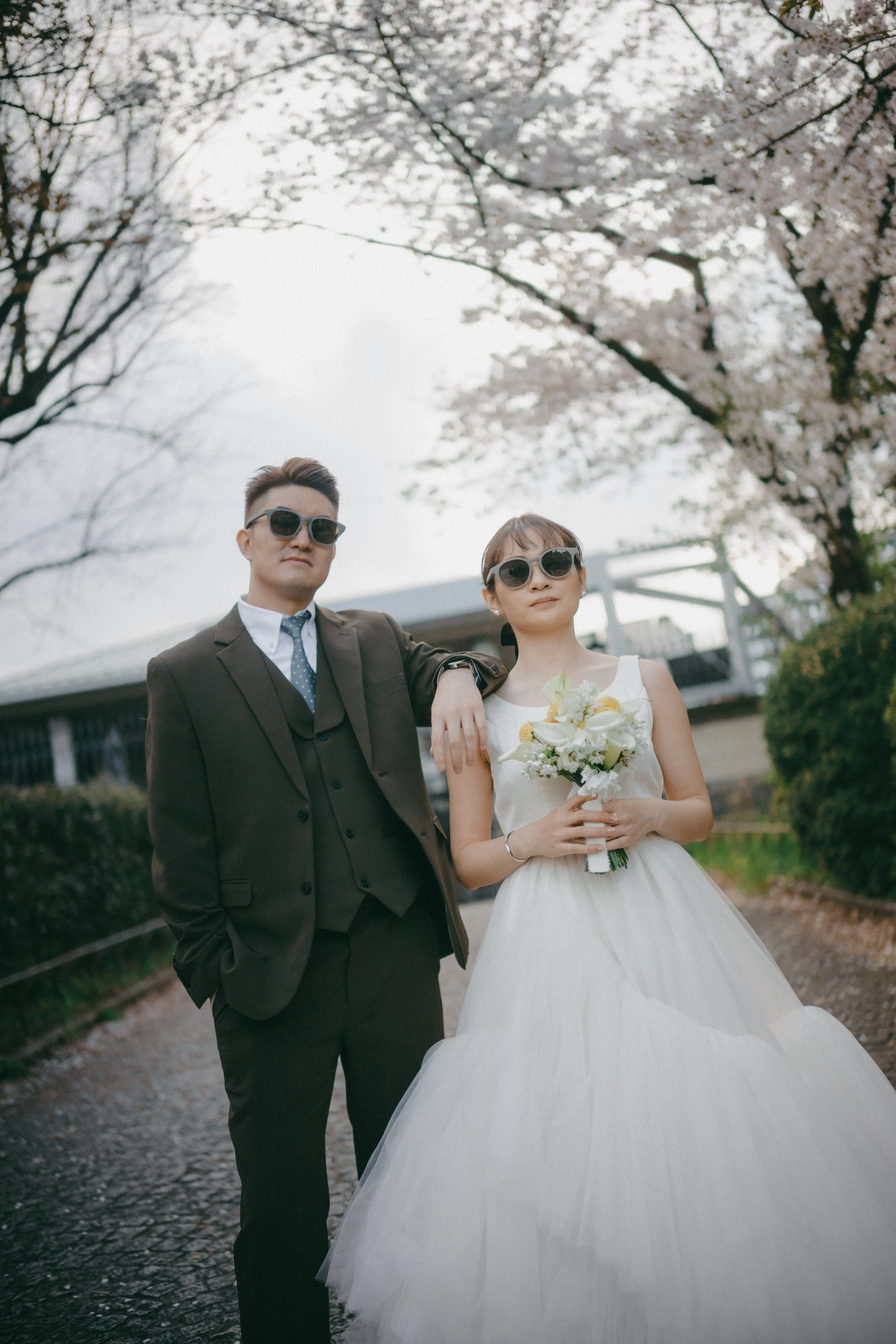 A man and woman in wedding attire, both wearing sunglasses, standing outdoors on a cobblestone path with blooming cherry blossom trees in the background. The woman is holding a bouquet of white flowers.