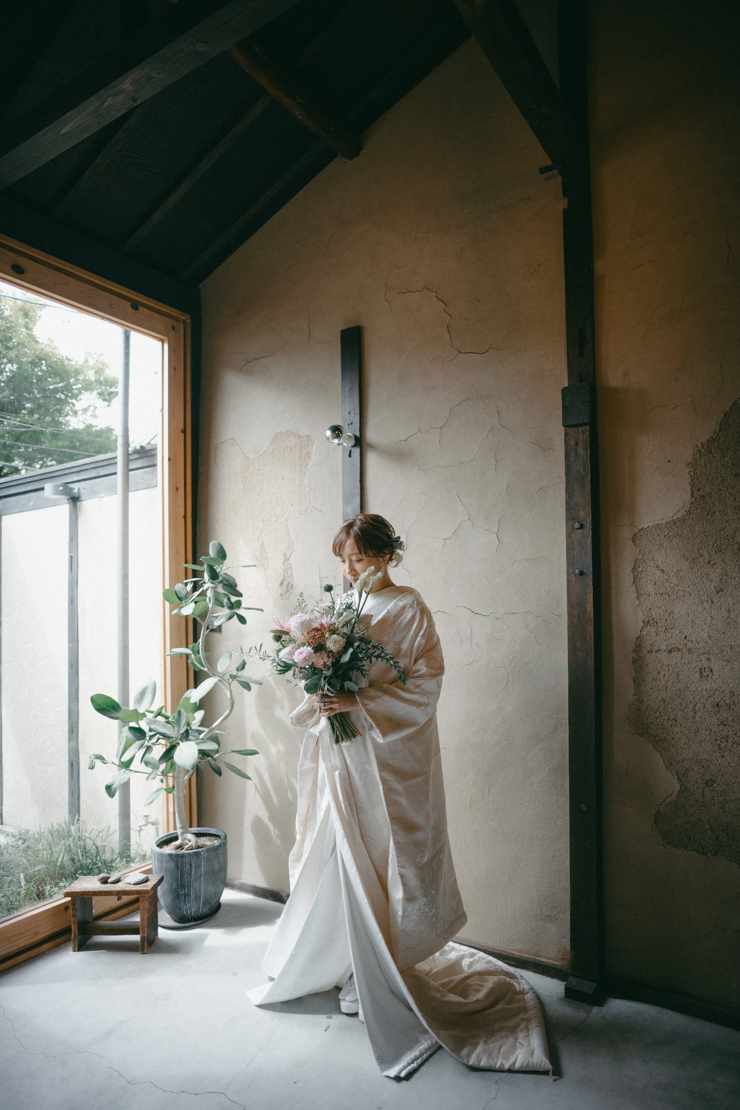 A woman in a wedding dress holding a bouquet of flowers, standing inside a room with a large window, an indoor plant in a pot, and a cracked wall.