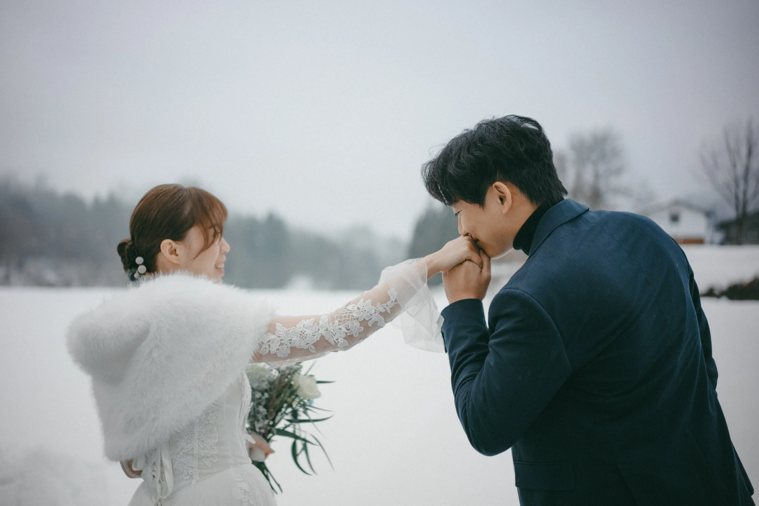 A man kneeling and kissing a woman's hand outdoors in a snowy landscape, with a woman in a white dress and shrug holding a bouquet of flowers.