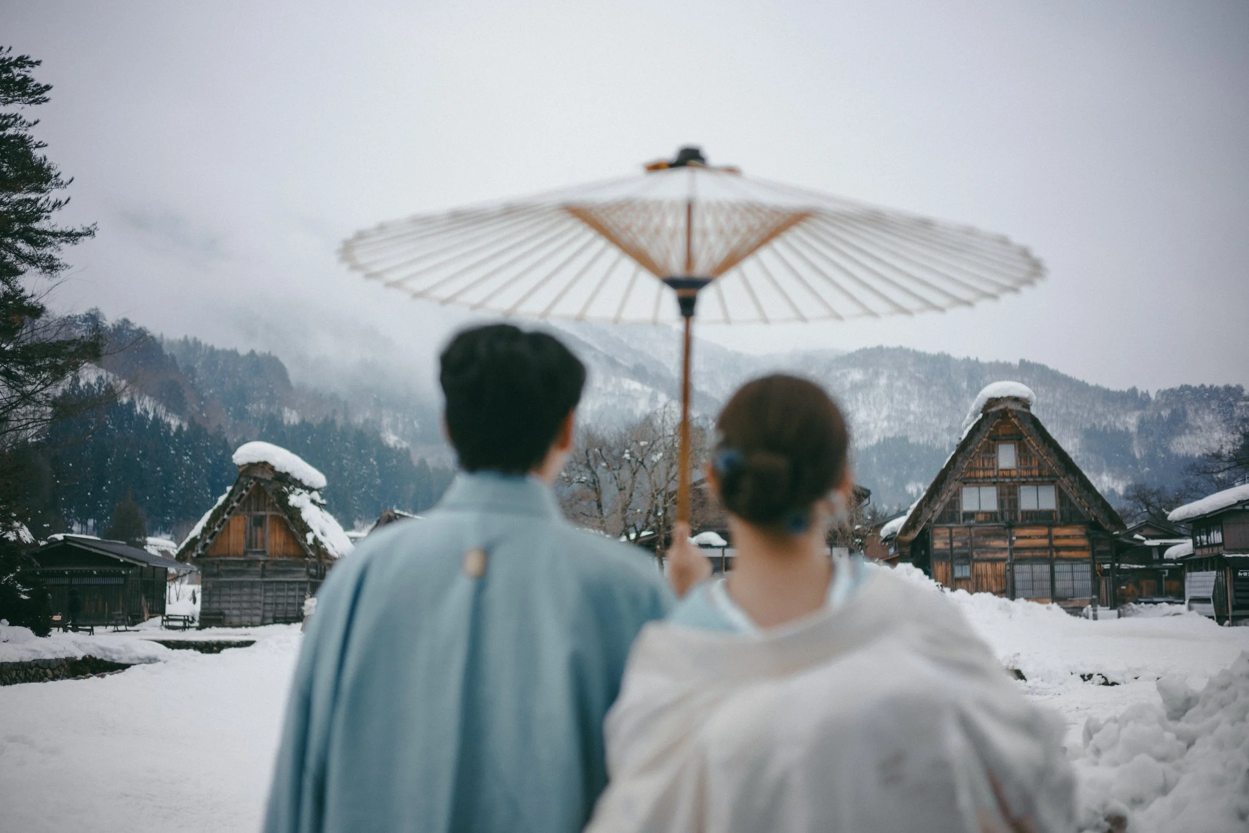 Two people dressed in traditional Japanese clothing, standing in a snowy village, holding a paper parasol, with wooden houses and snow-covered mountains in the background.