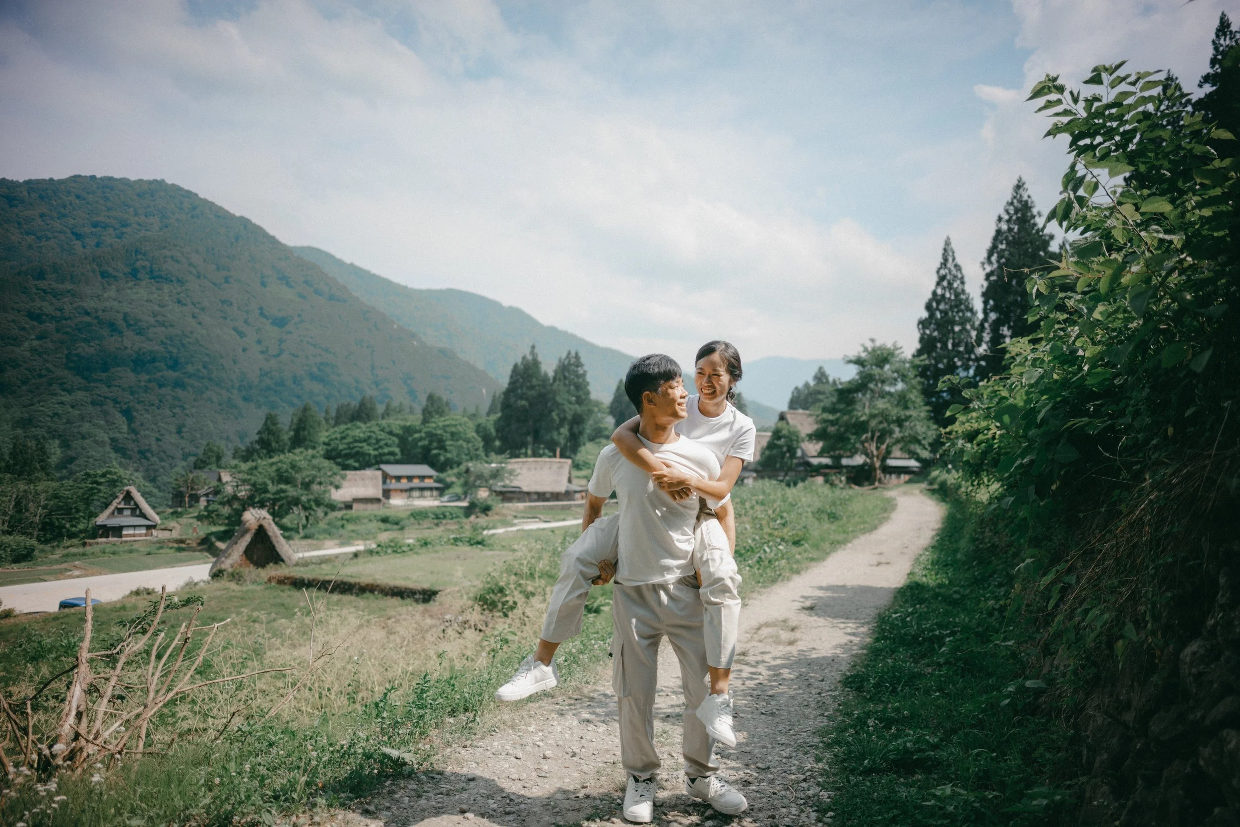 A couple playing and laughing on a rural dirt path in a green mountain village, surrounded by trees and traditional houses.