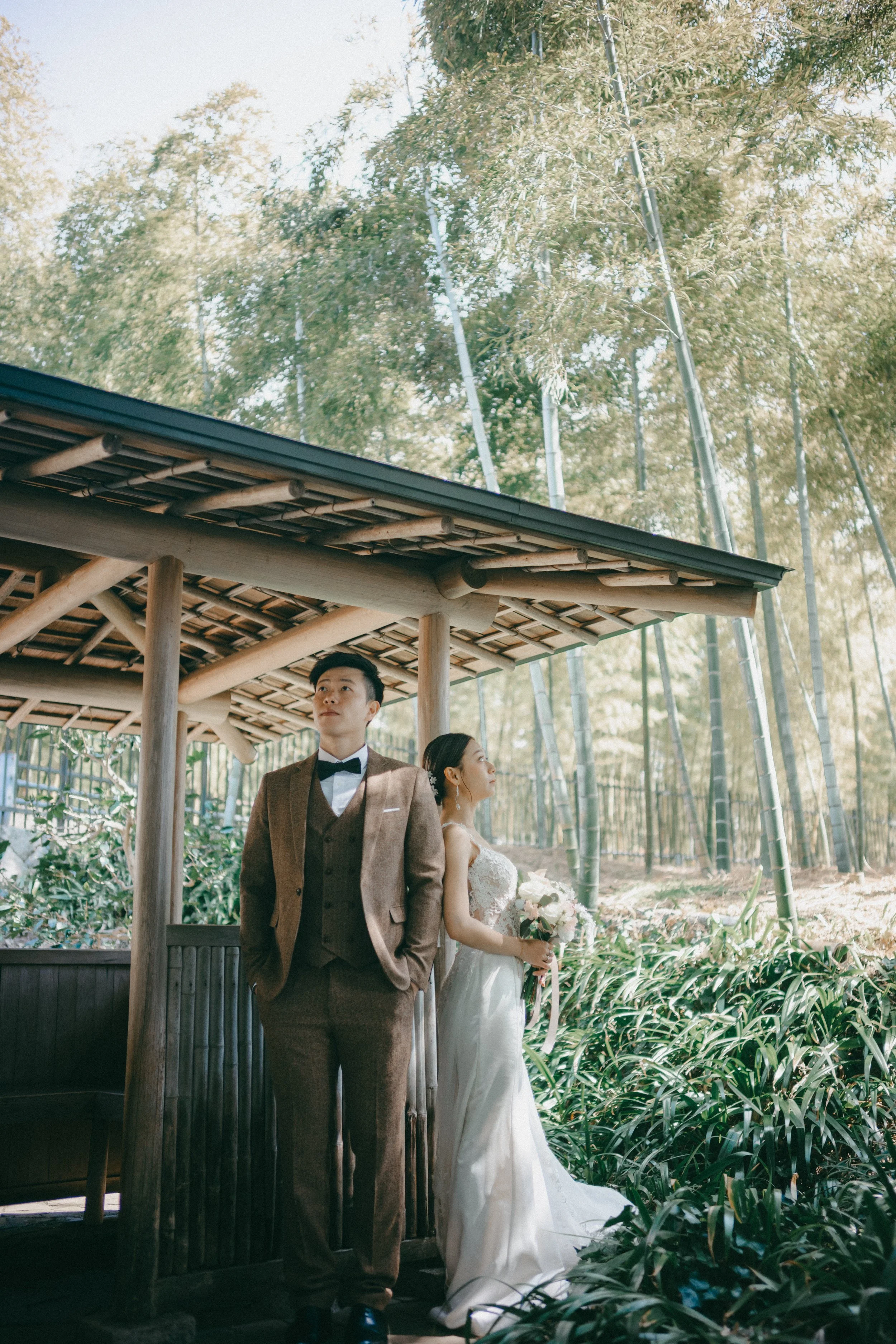A bride in a white wedding gown holding a bouquet stands near a groom in a brown suit with a bow tie, outdoors in a lush garden with bamboo trees.