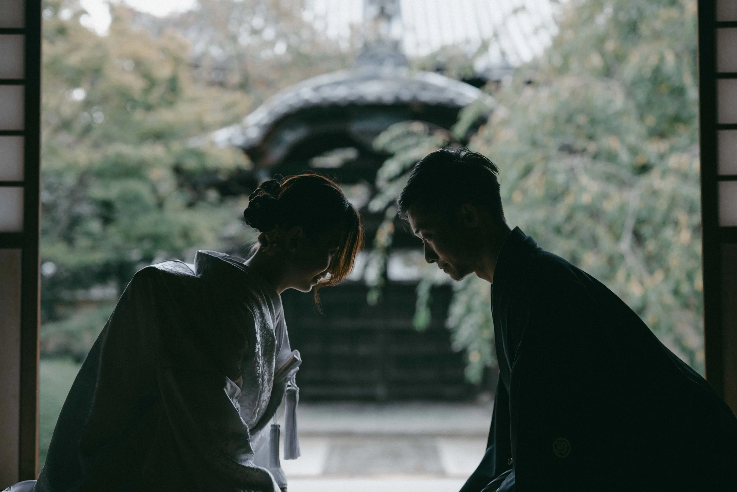 Silhouettes of a man and woman in traditional Japanese attire bowing to each other inside a Japanese temple with a garden in the background.