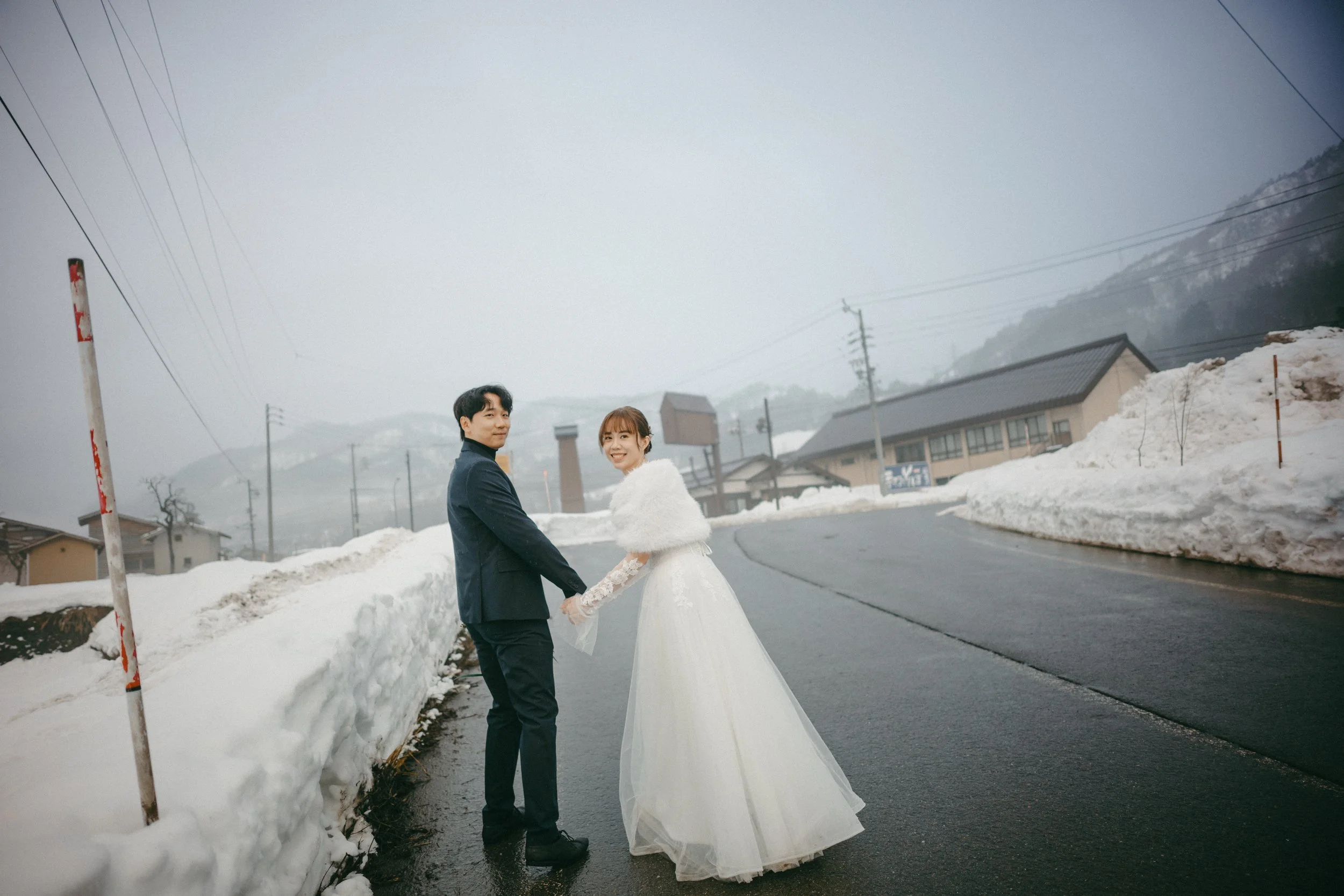 A couple in wedding attire holding hands on a snowy mountain road, with snow banks and foggy mountains in the background.