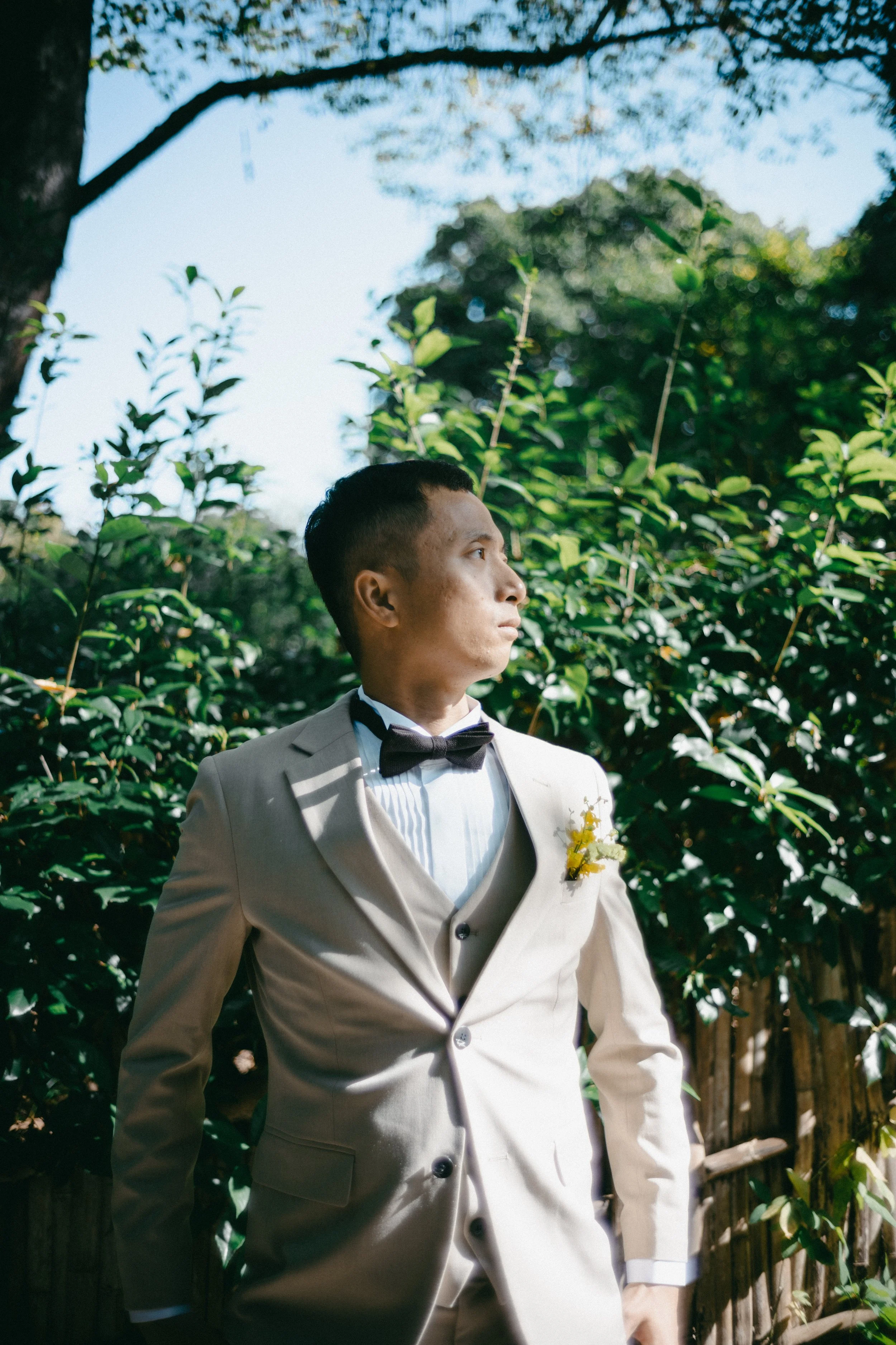 A man in a beige suit, white dress shirt, and black bow tie standing outdoors among green foliage, looking to the right.