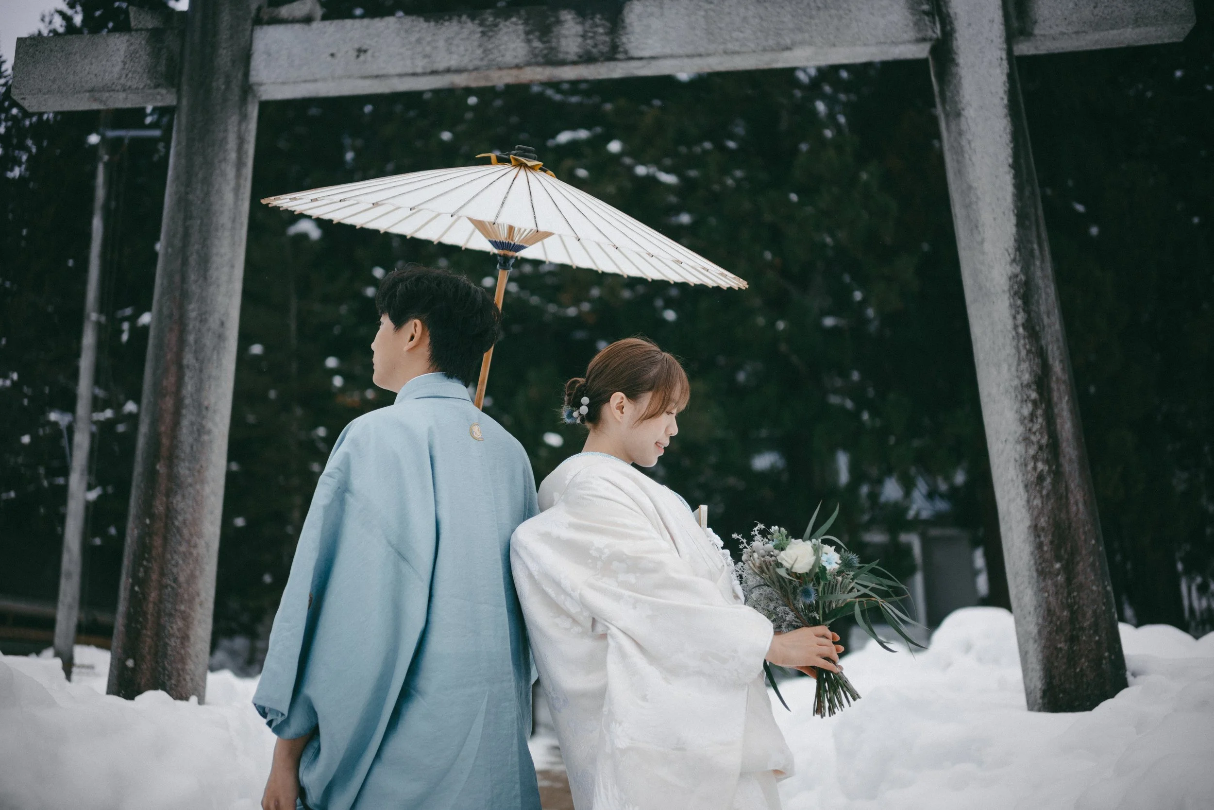 A couple dressed in traditional Japanese wedding attire standing under a wooden torii gate in the snow, with the groom holding a white parasol and the bride holding a bouquet of flowers.