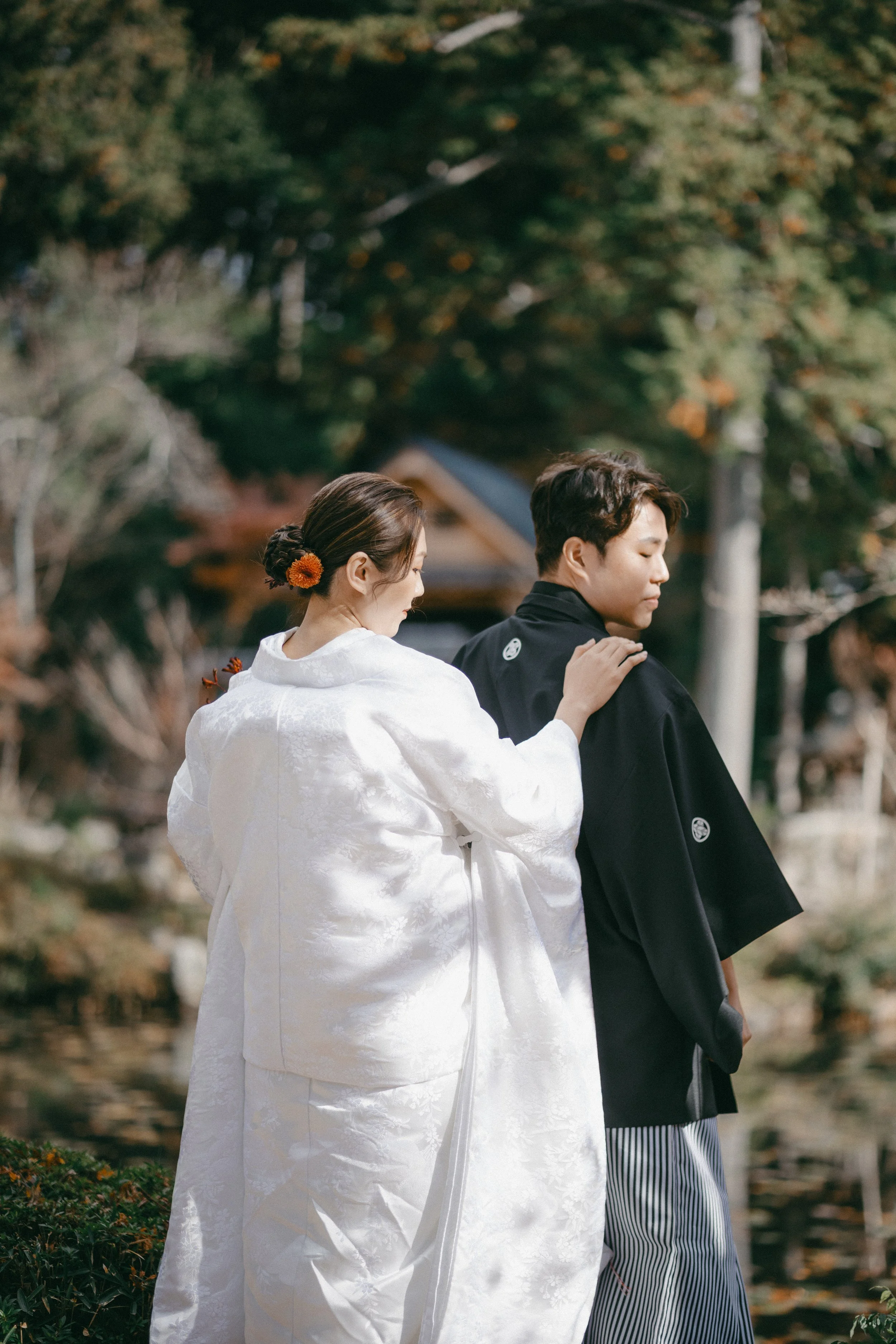 A woman in a white traditional Japanese wedding kimono gently touches a man in a black formal kimono, with both standing outdoors near a body of water and trees.
