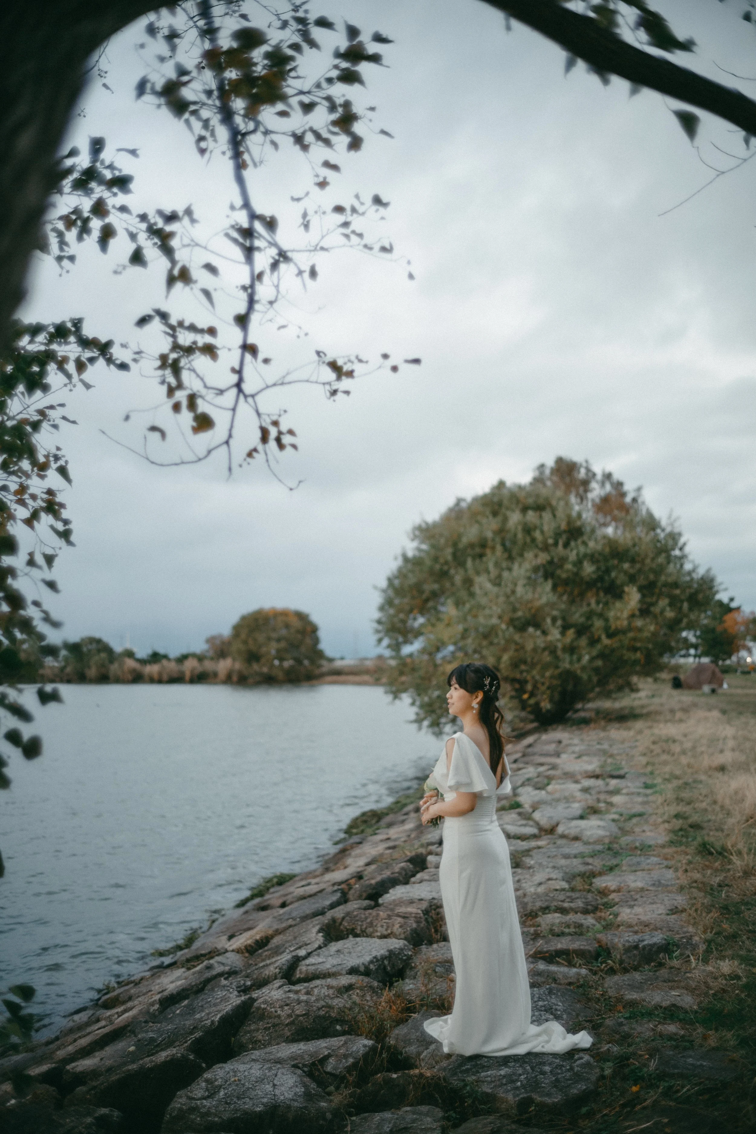 A woman in a white dress standing on a rocky riverside path, gazing into the distance with trees and water in the background.
