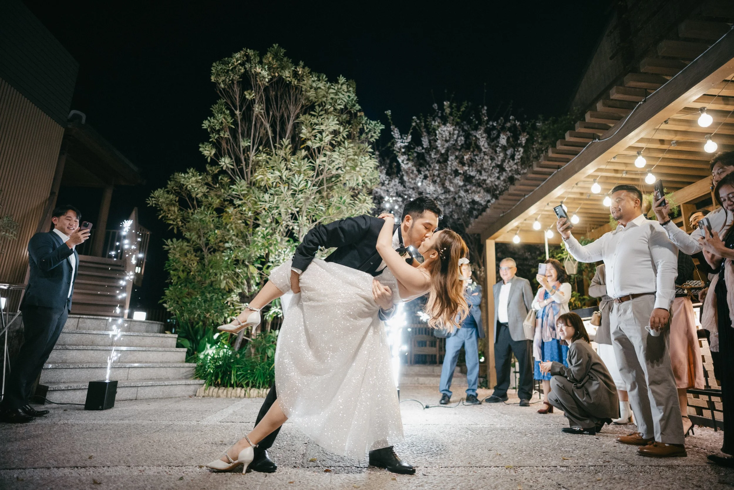 A bride and groom sharing a kiss during their wedding reception at night, surrounded by guests taking photos.
