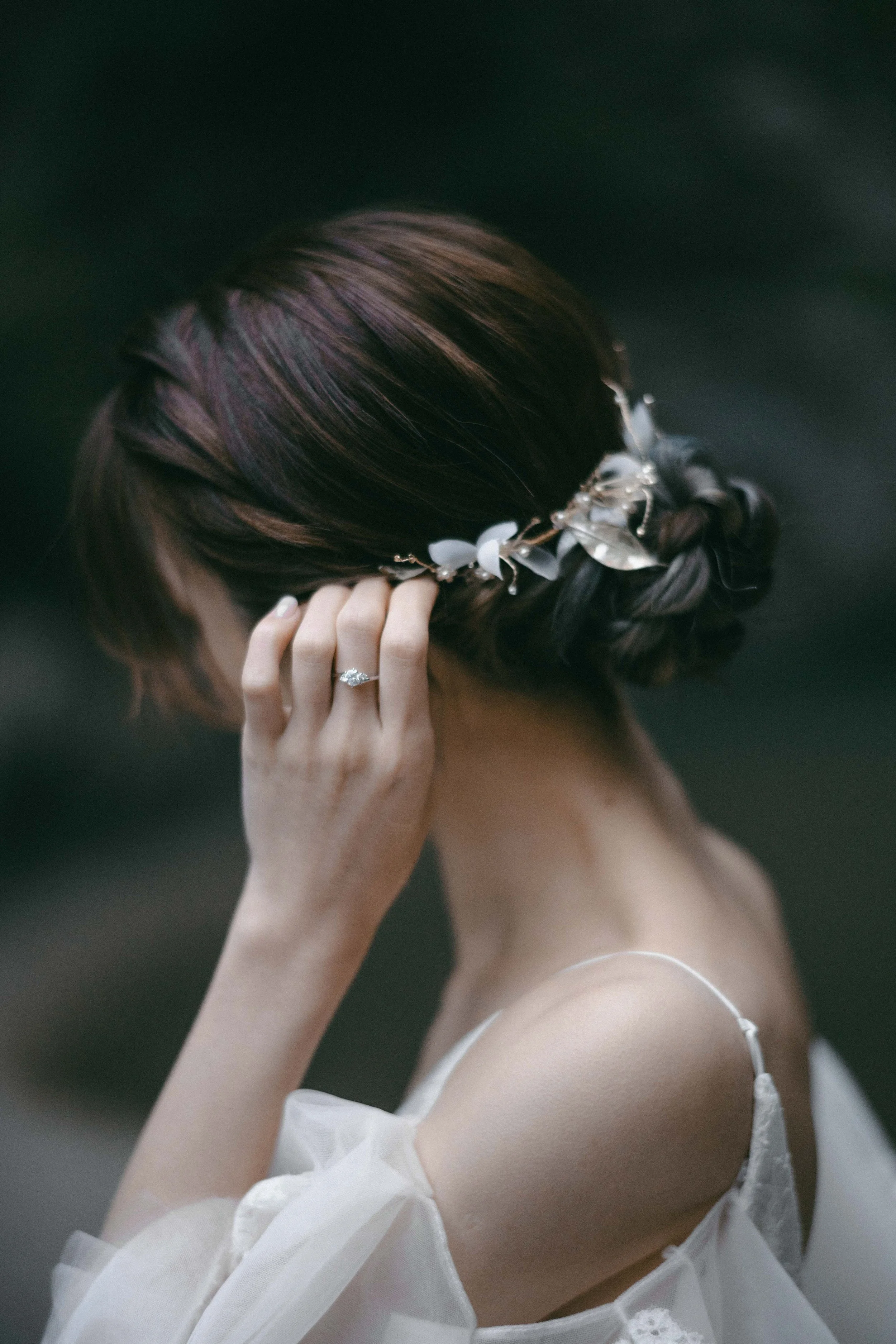 A woman with brown hair styled in an updo, wearing a decorative headpiece, and a white dress with thin straps, holding her hand near her face showing a ring on her finger. The background is dark and blurred.