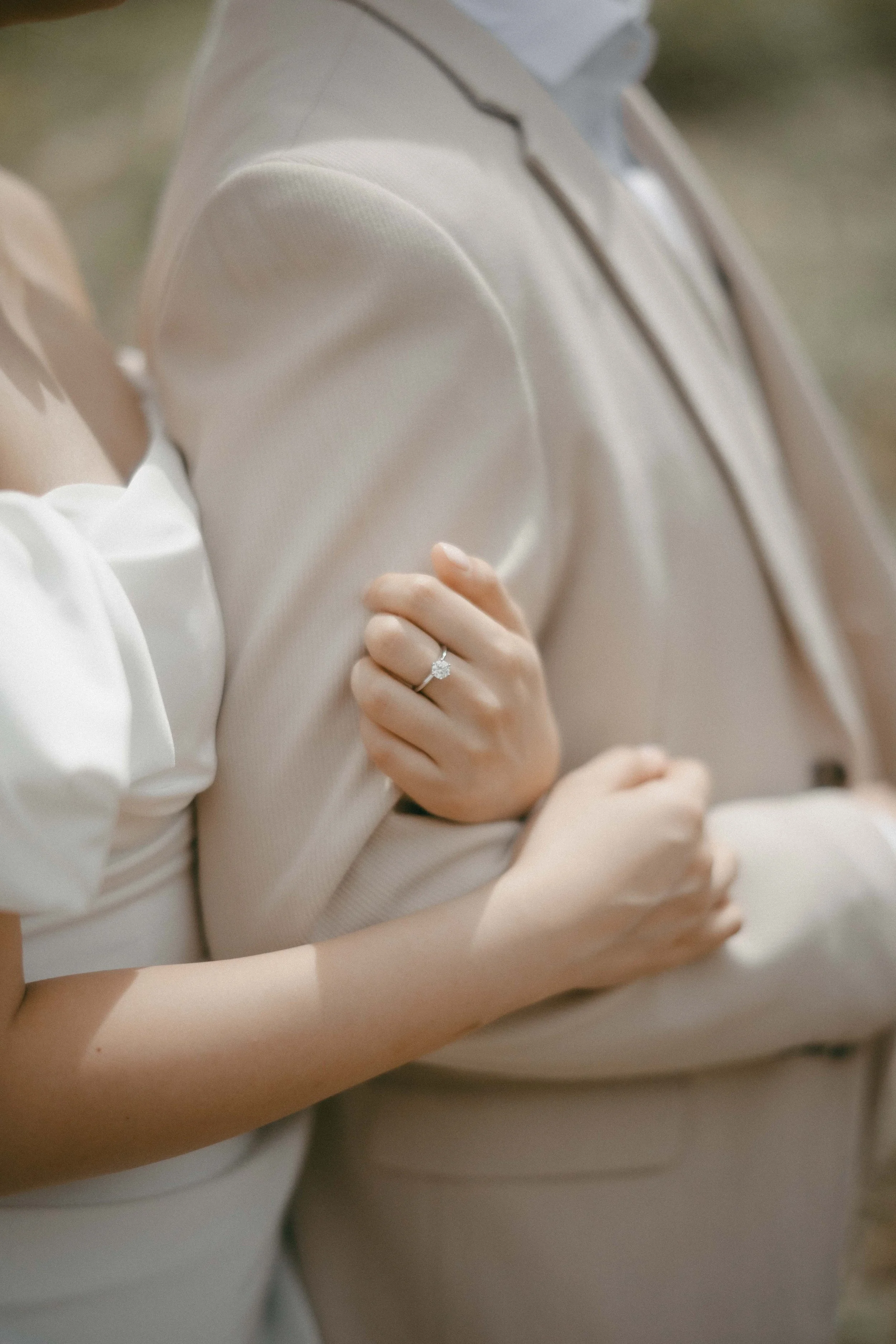 Close-up of a woman wearing a beige suit and an engagement ring on her finger, with her arm gently holding someone else's arm.