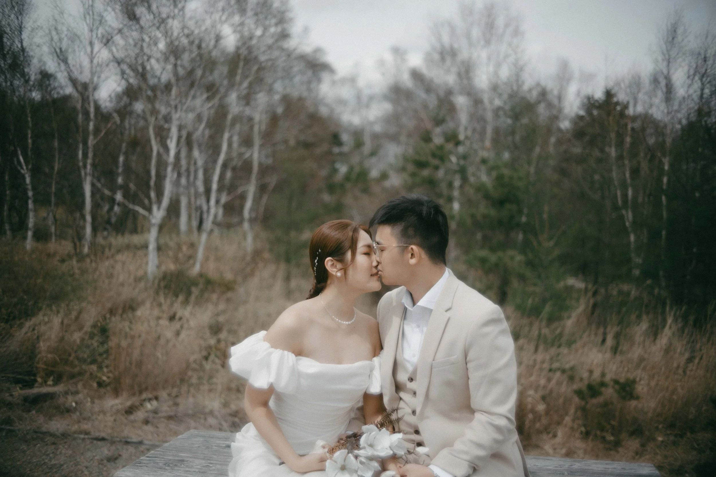 A bride and groom sitting on a wooden plank in a natural outdoor setting with trees, kissing with their eyes closed, holding a bouquet of flowers.