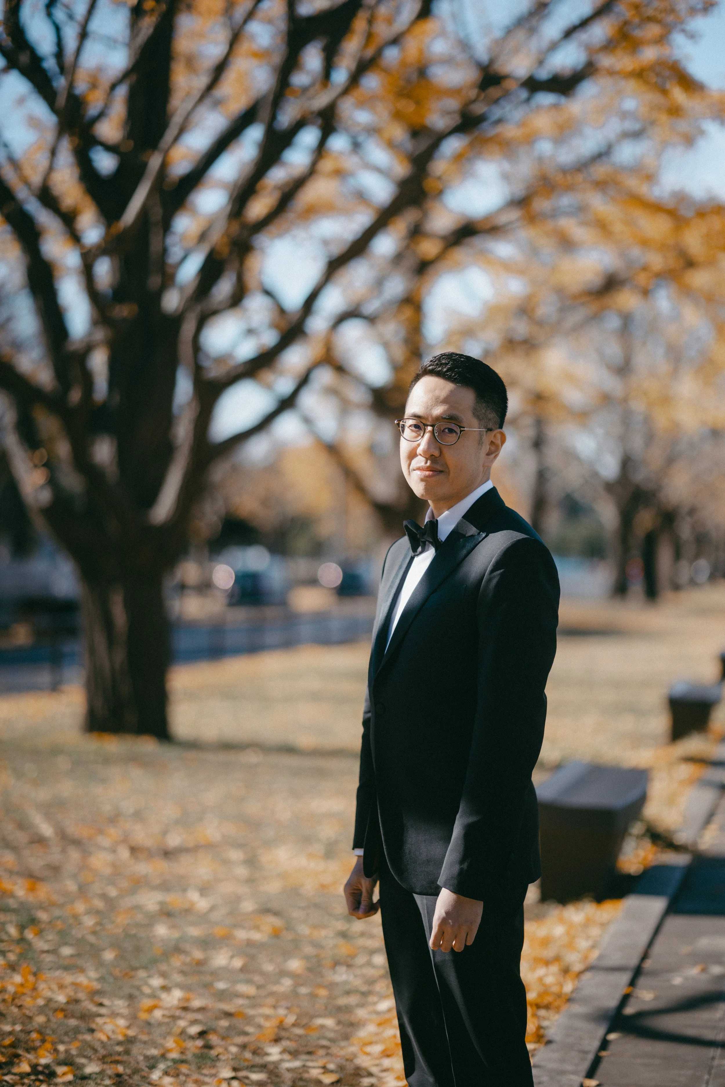 A man in a black tuxedo with a bow tie standing outdoors during autumn, with yellow-orange leaves on the trees and fallen leaves on the ground.