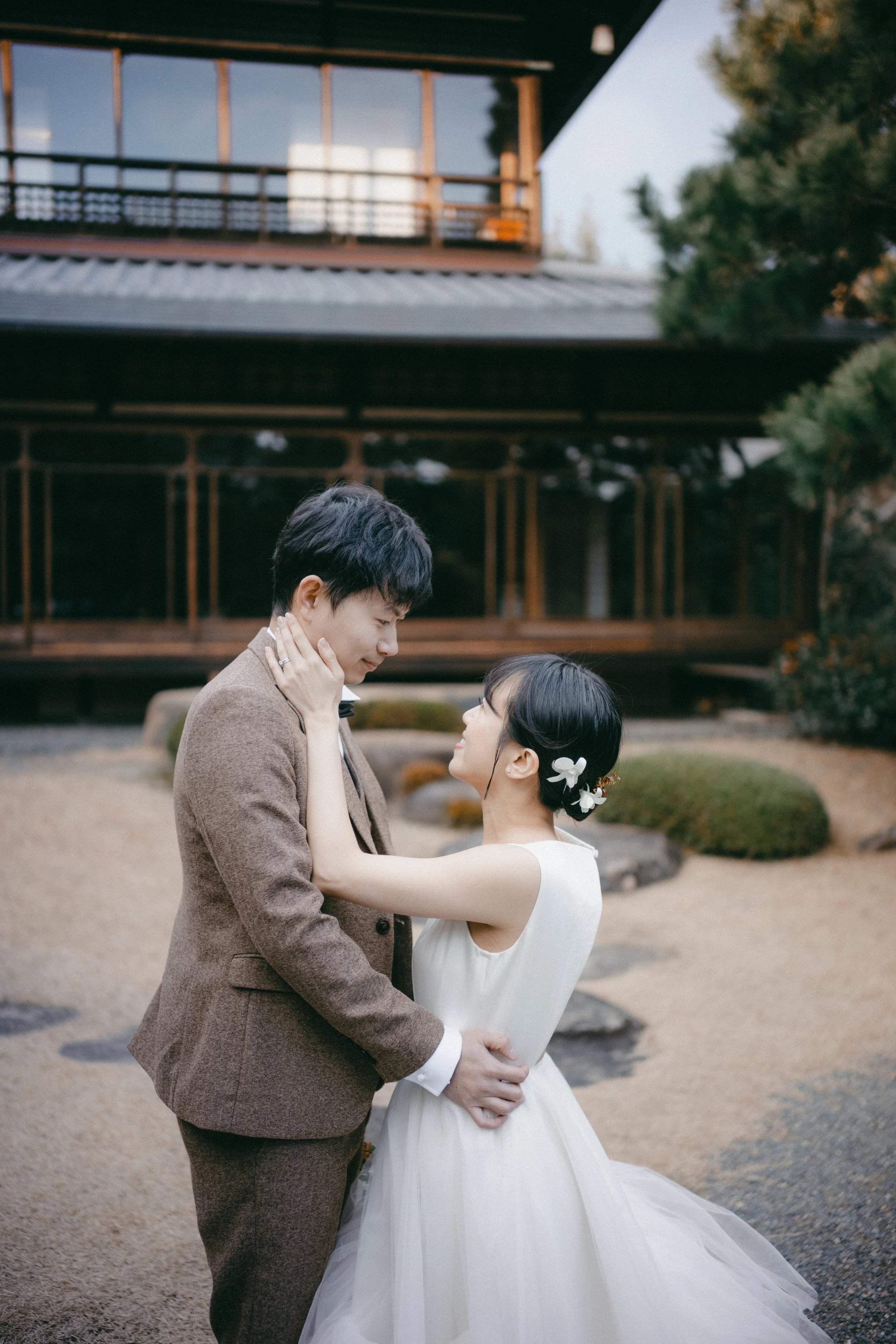 A couple dressed in wedding attire standing close together outdoors, gazing into each other's eyes, with a traditional Japanese-style building in the background.