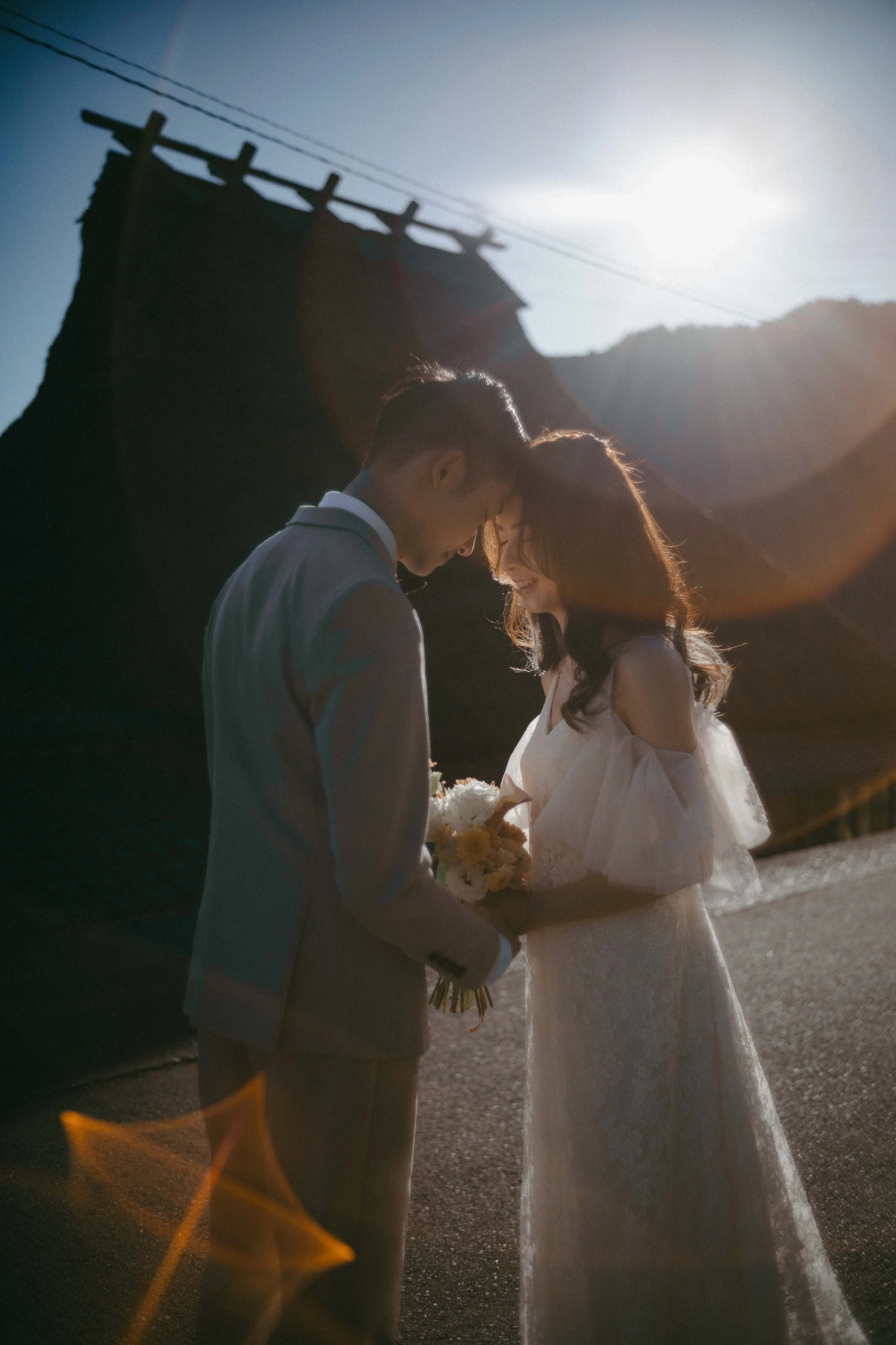 A couple dressed in wedding attire standing close together outdoors at sunset, smiling, with mountains and a building in the background.