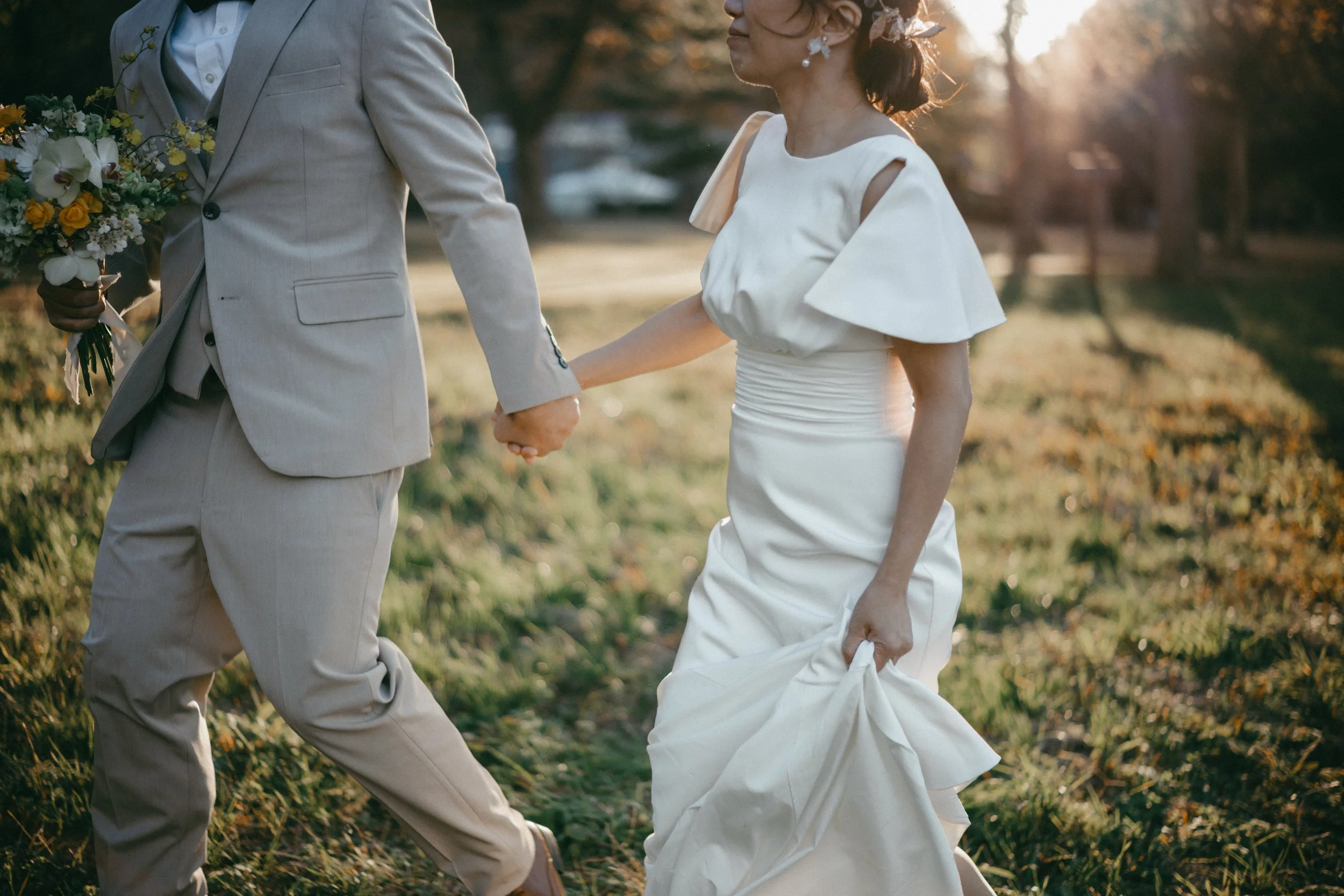 A couple holding hands, walking outdoors during sunset, with the woman wearing a white dress and the man in a light-colored suit. The woman is lifting her dress slightly, and the man is holding a bouquet of flowers.