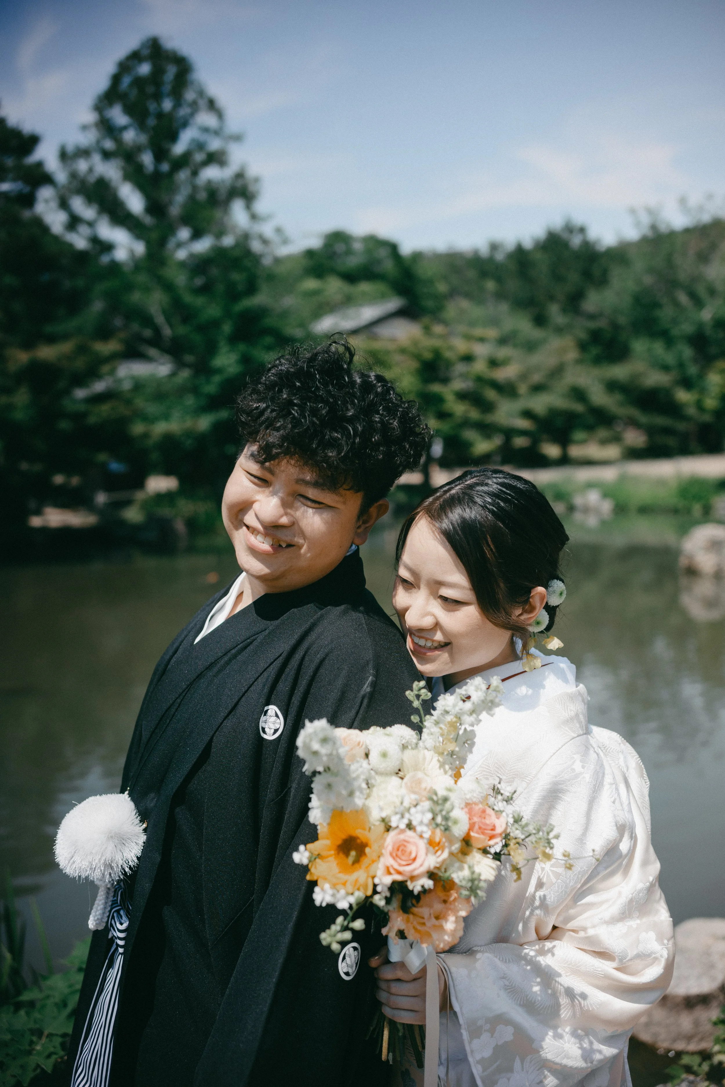 A happy couple in traditional Japanese wedding attire outdoors near a pond, with trees and a blue sky in the background.