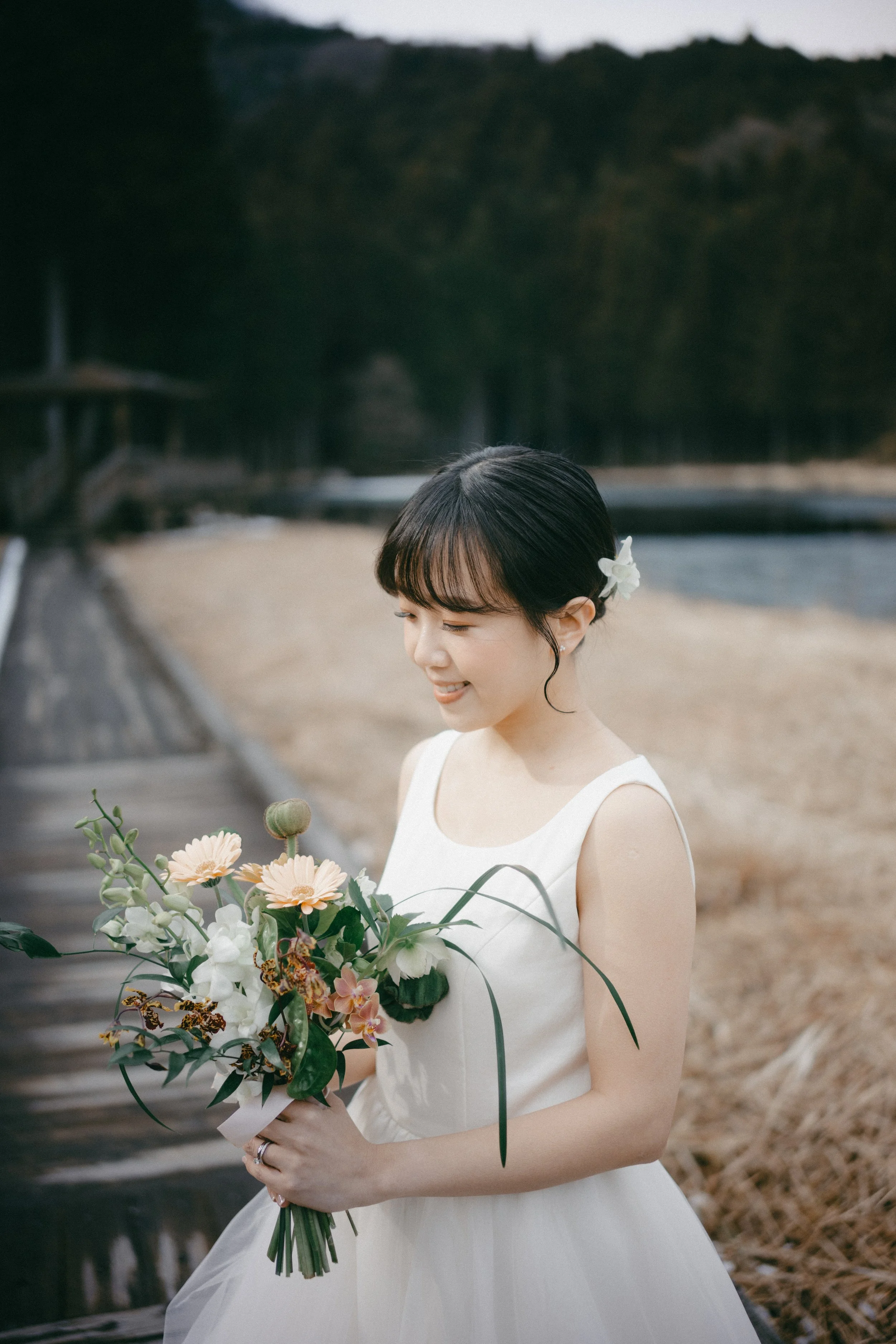 A woman in a white dress holding a bouquet of flowers outdoors near a body of water with trees and a mountain in the background.