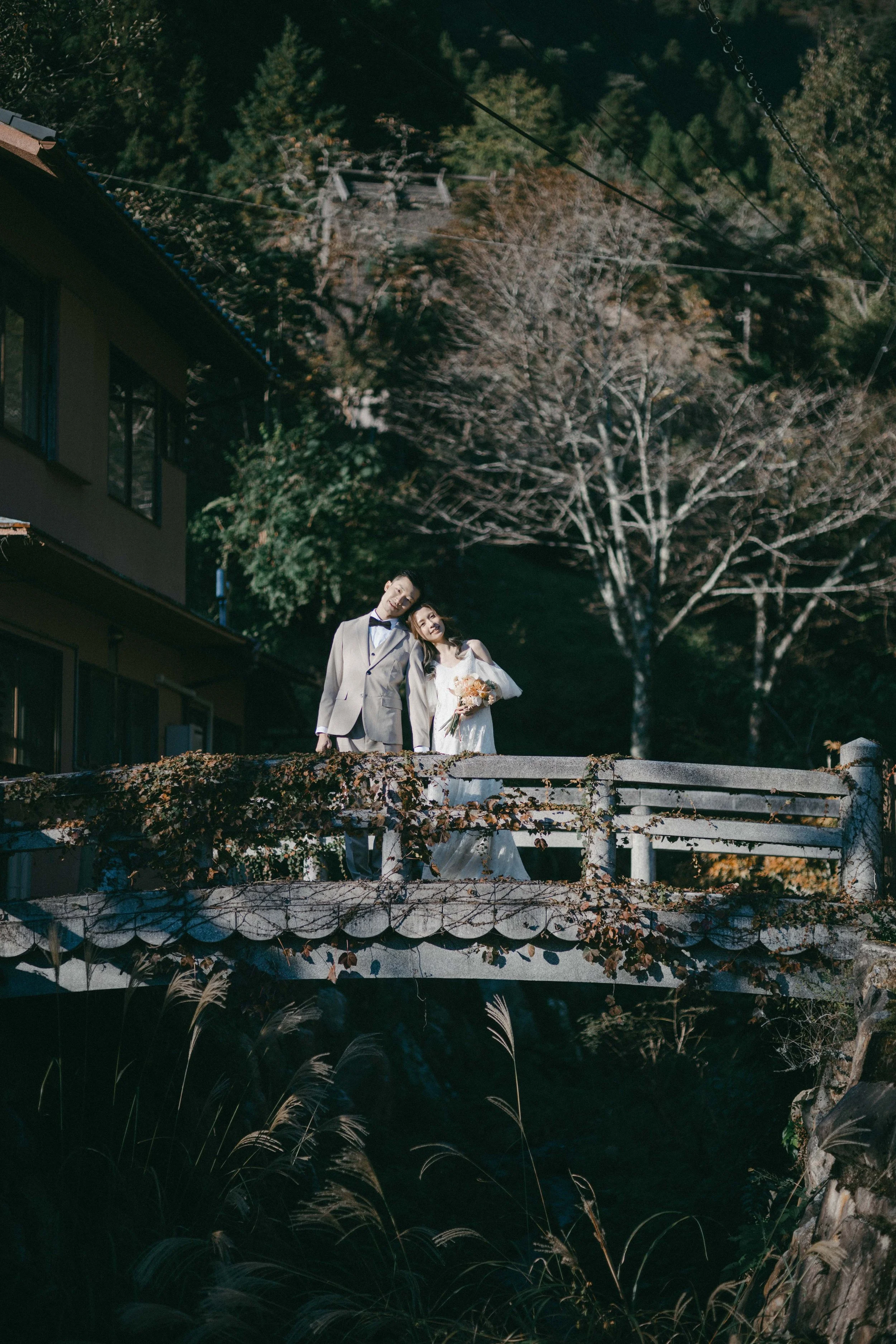 A couple dressed in wedding attire standing on a small bridge outdoors, holding a bouquet of flowers, with trees and a building in the background.