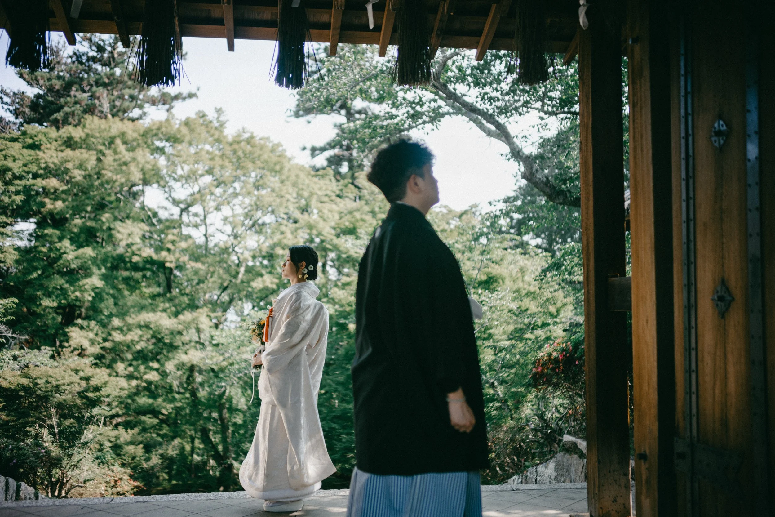 A man and a woman dressed in traditional Japanese wedding attire standing outdoors under a wooden structure with green trees in the background.