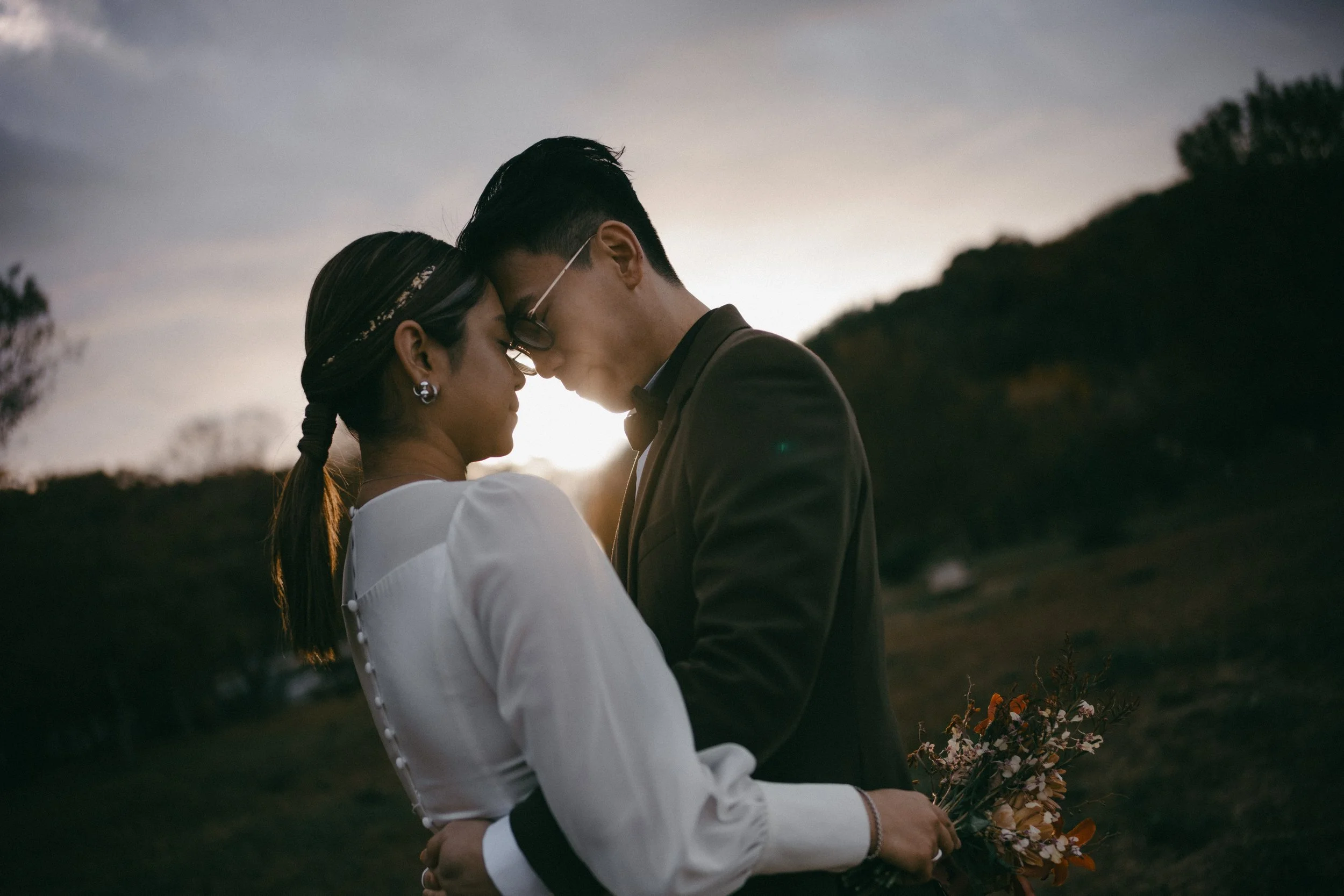 A couple embraces outdoors at sunset, with noses touching and holding a bouquet.