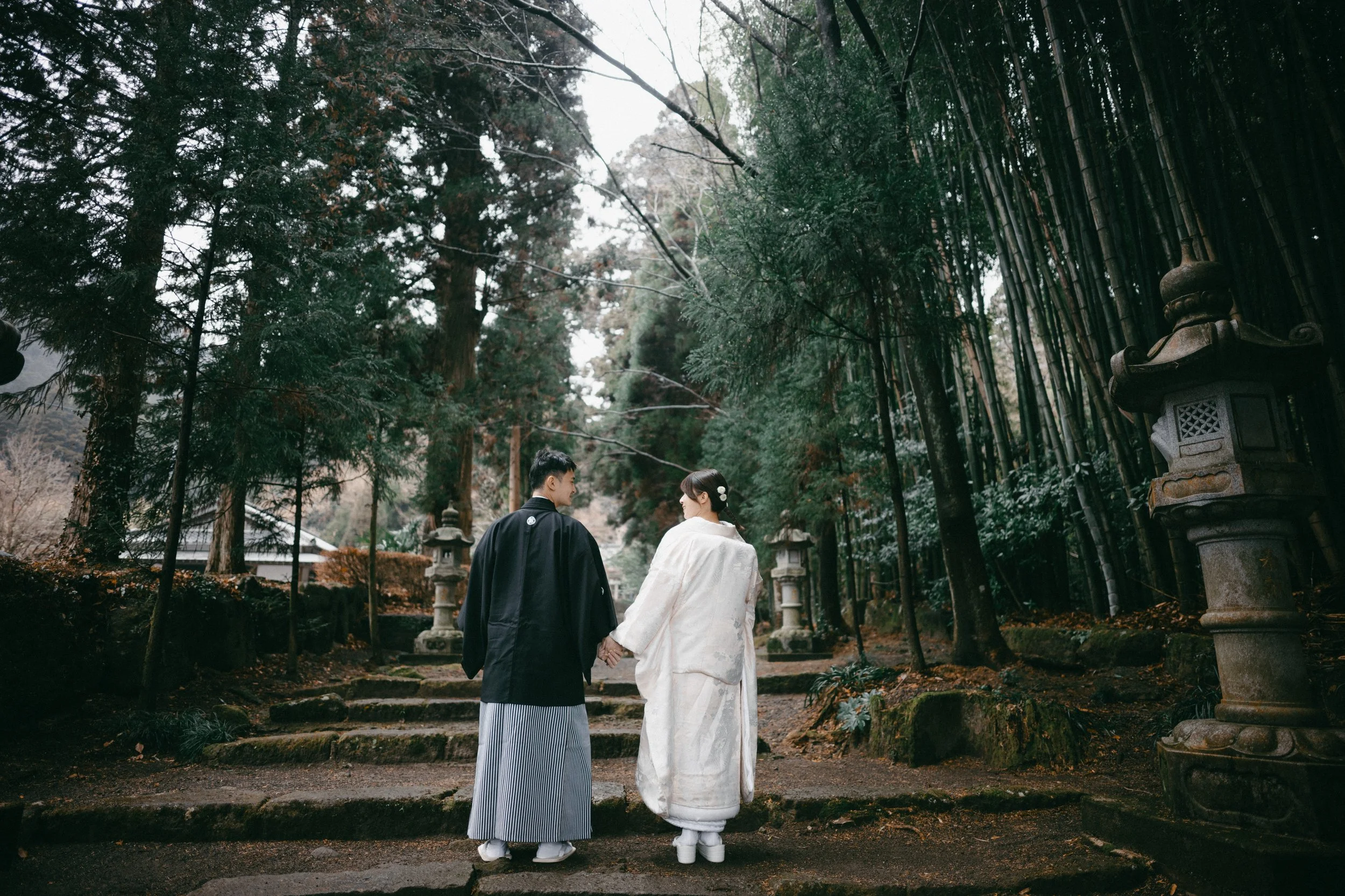 A couple dressed in traditional Japanese wedding attire walking hand in hand on stone steps in a lush forest with tall trees and stone lanterns.
