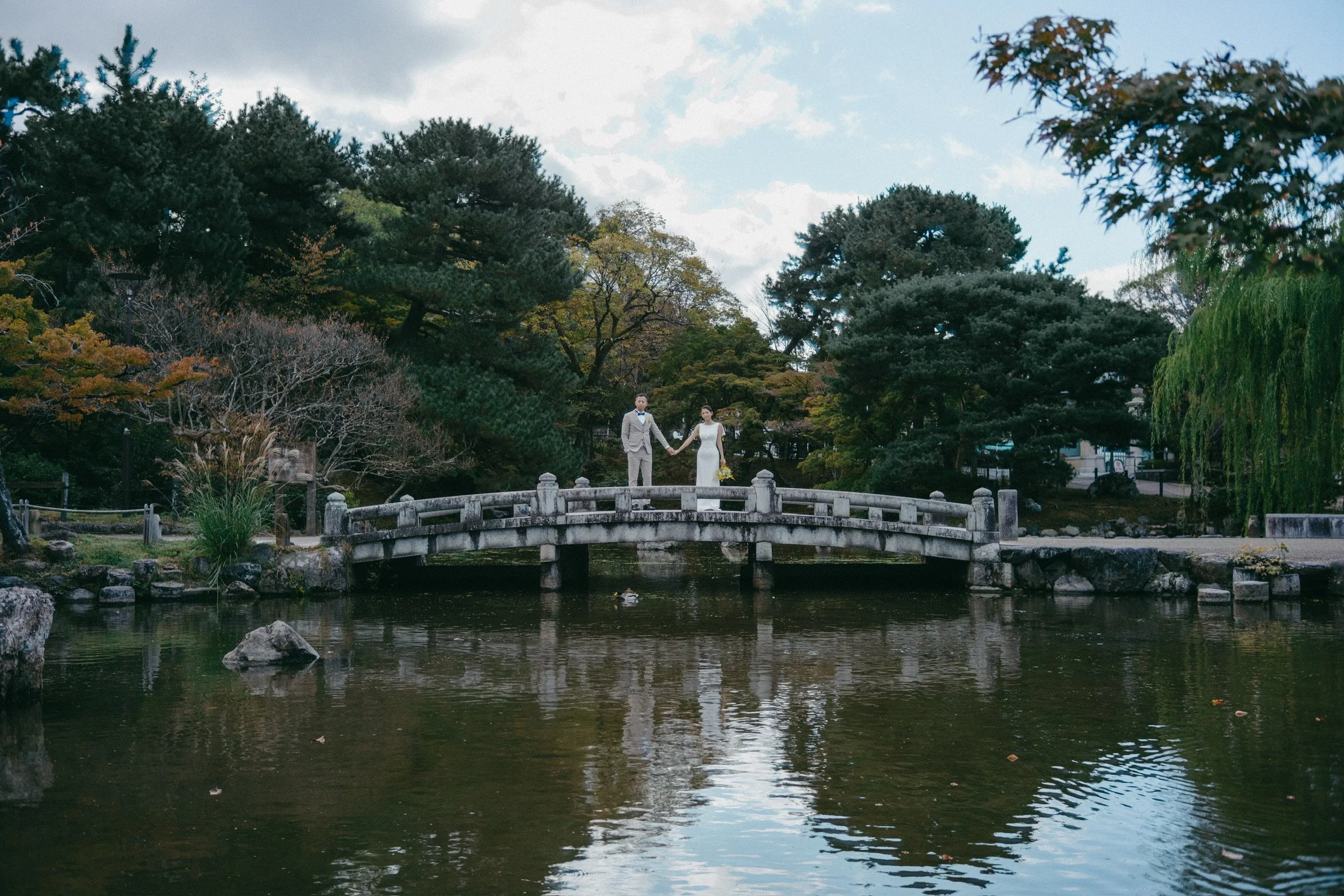 A bride and groom holding hands standing on a small stone bridge over a pond in a park with trees and cloudy sky in the background.