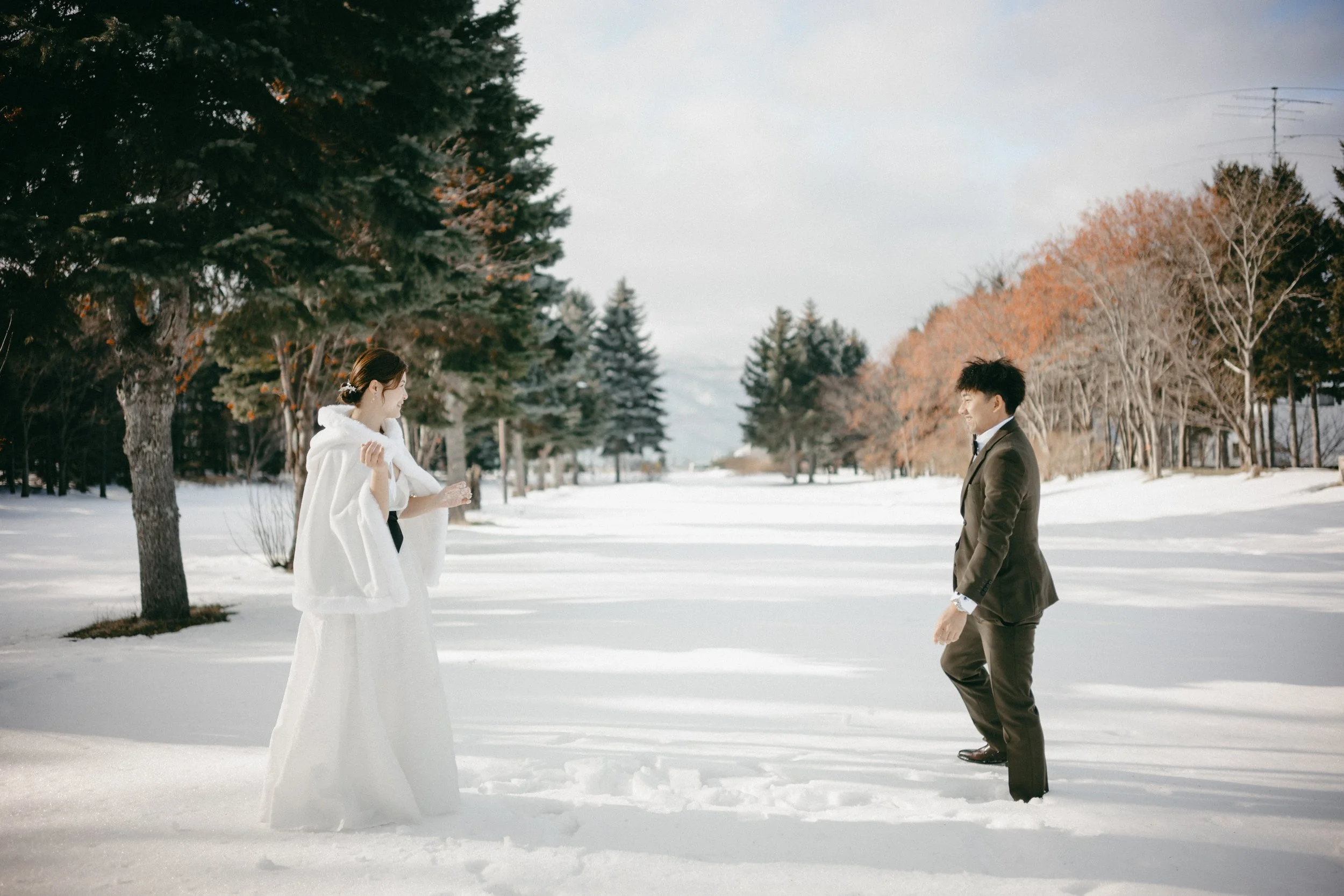 A man in a black suit and a woman in a white wedding dress standing apart in a snow-covered landscape with trees and an overcast sky.