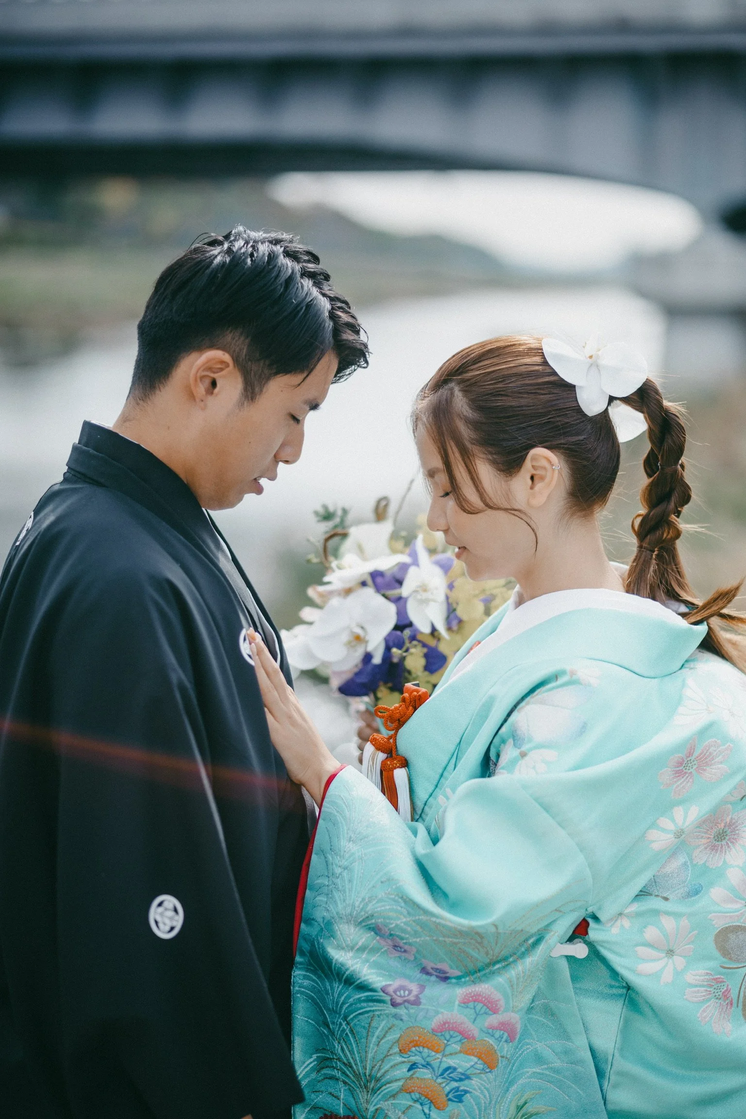 A man and a woman in traditional Japanese attire sharing an intimate moment outdoors near a river, with a bridge in the background. The woman wears a light blue kimono with floral designs and has a white flower in her hair. The man wears a dark kimon
