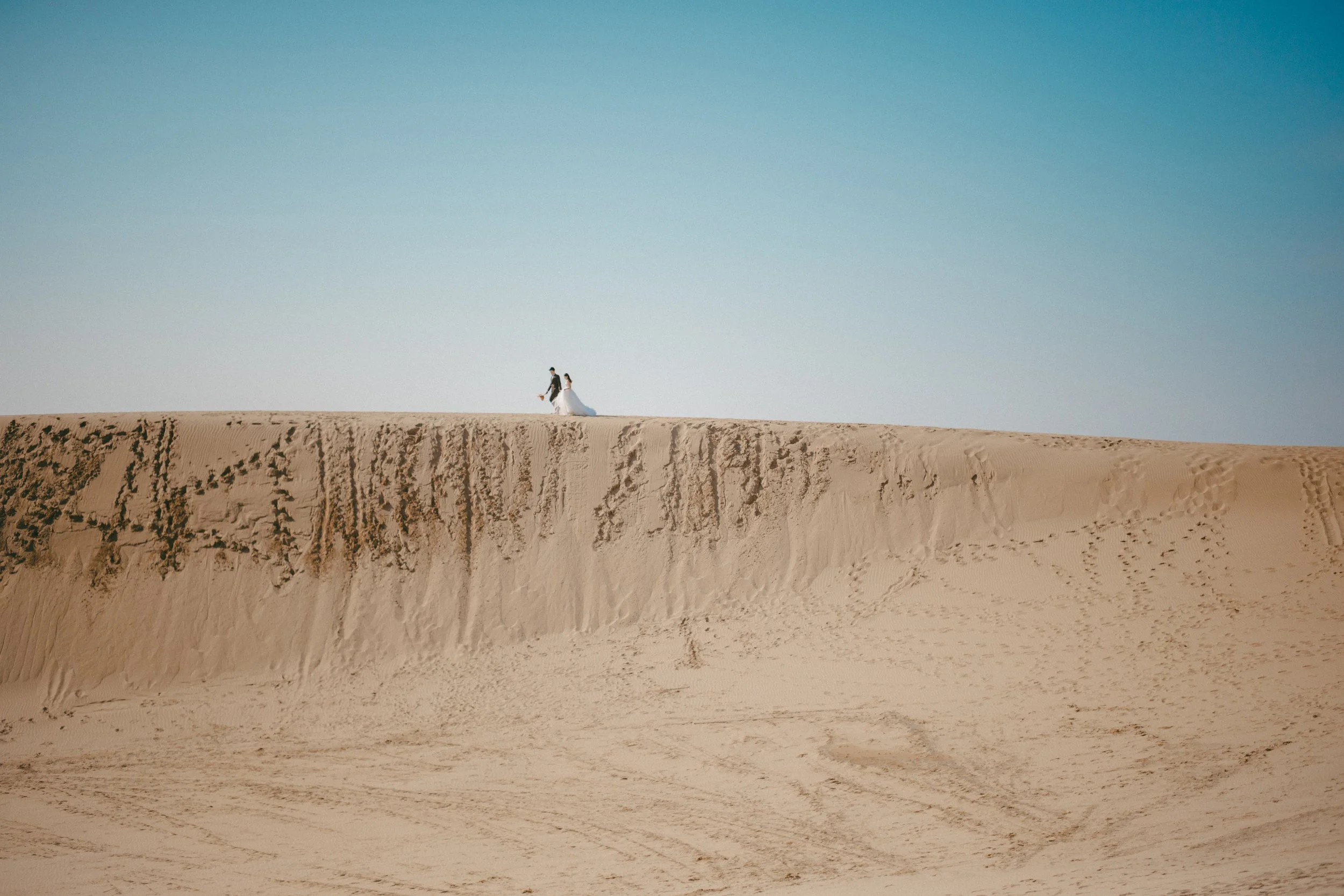 A bride and groom walk on a sand dune in a desert, with clear blue sky above.