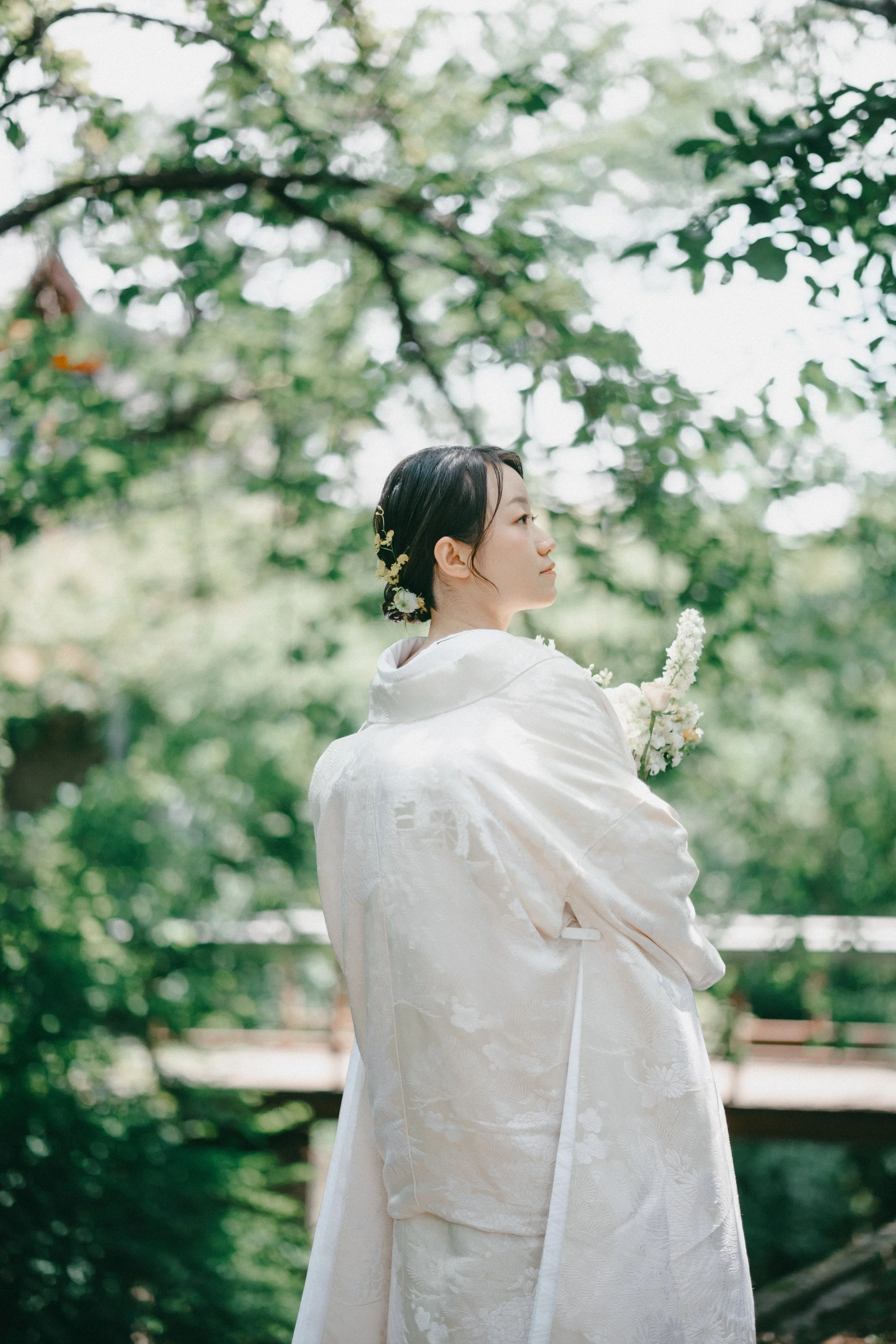 A woman in traditional Japanese wedding attire holding a bouquet of flowers, standing outdoors among green trees.