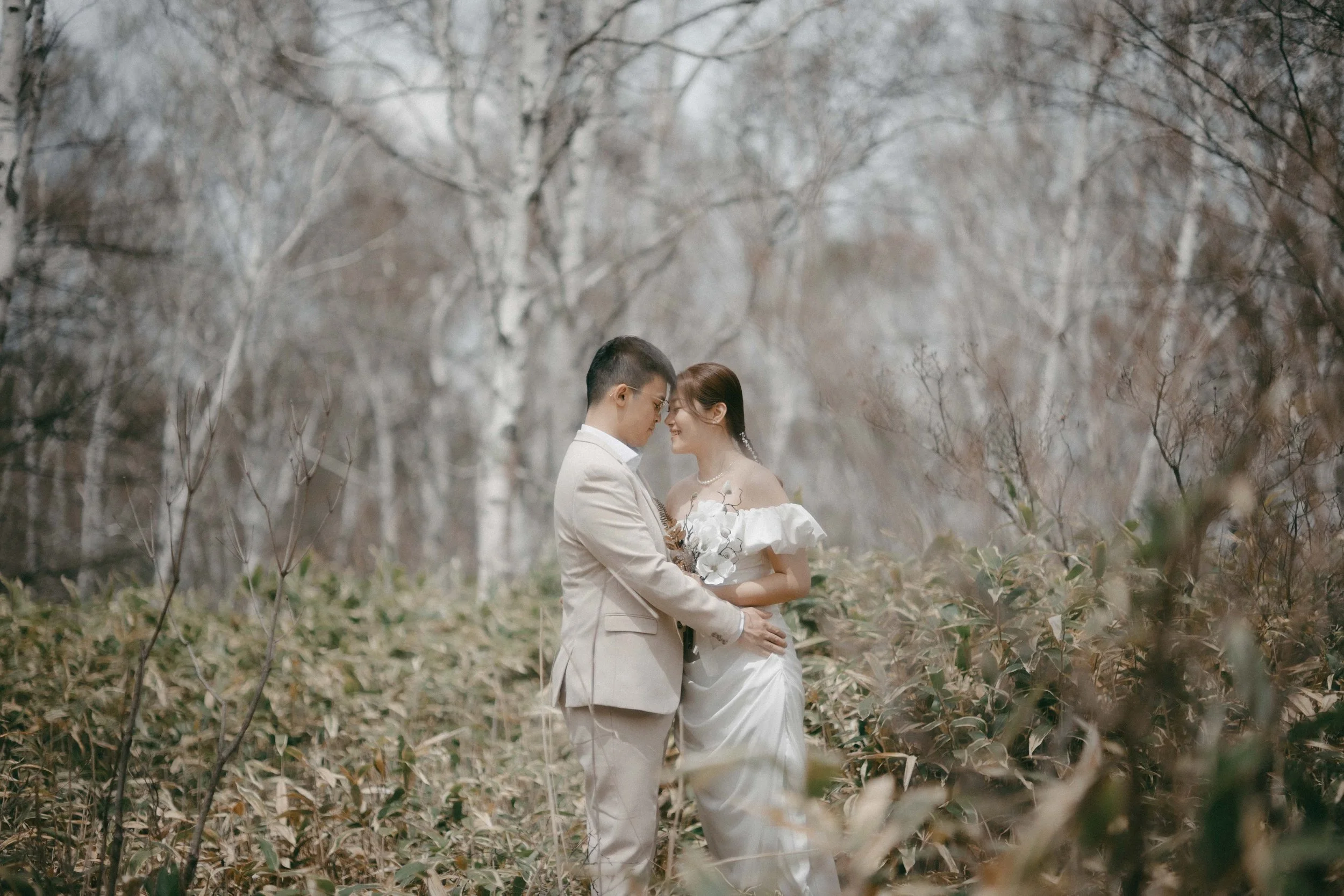 A couple in wedding attire sharing an intimate moment in a forest with bare trees and fallen leaves.