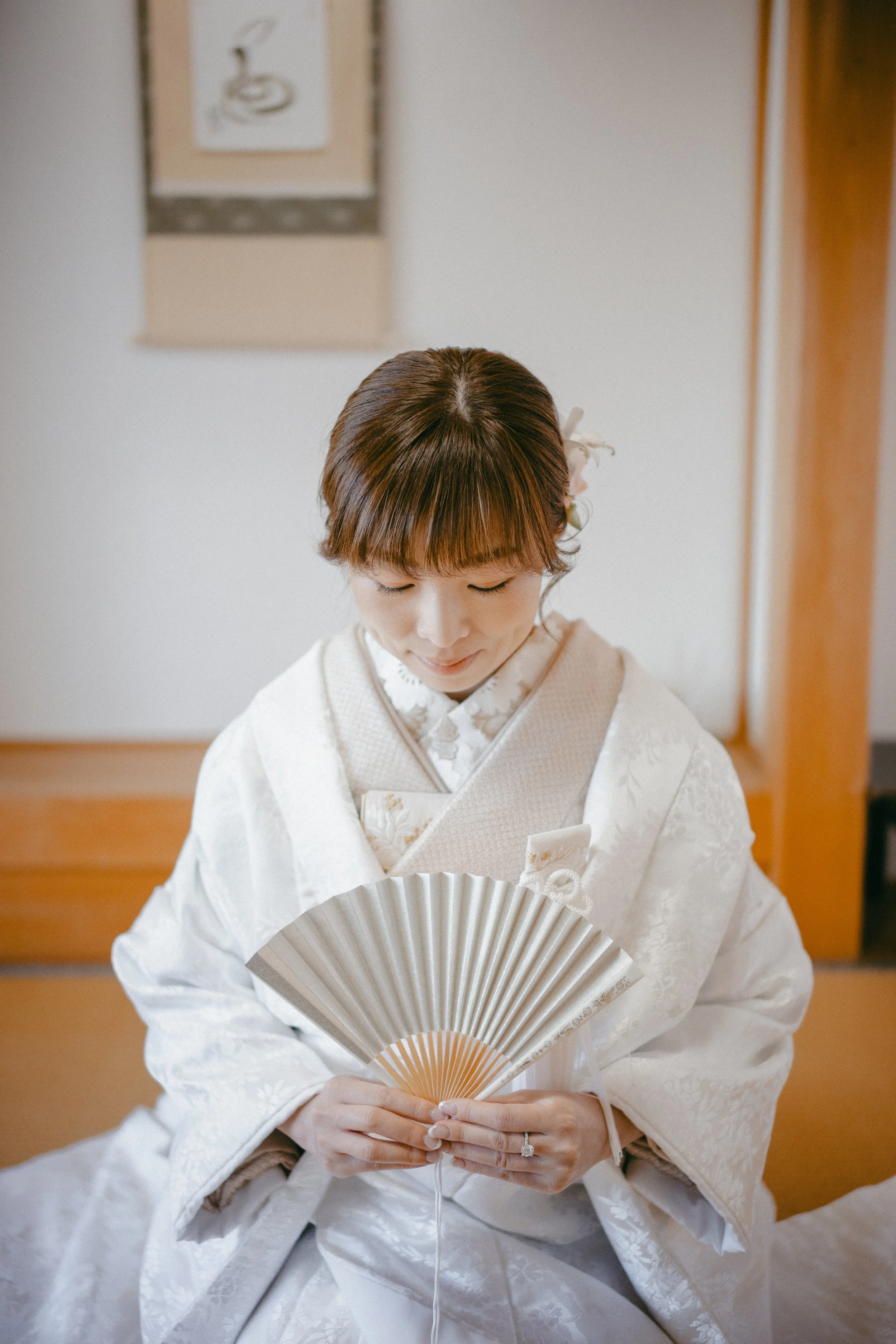 A woman dressed in traditional Japanese attire holding a folding fan, sitting in a room with wooden elements and artwork on the wall.