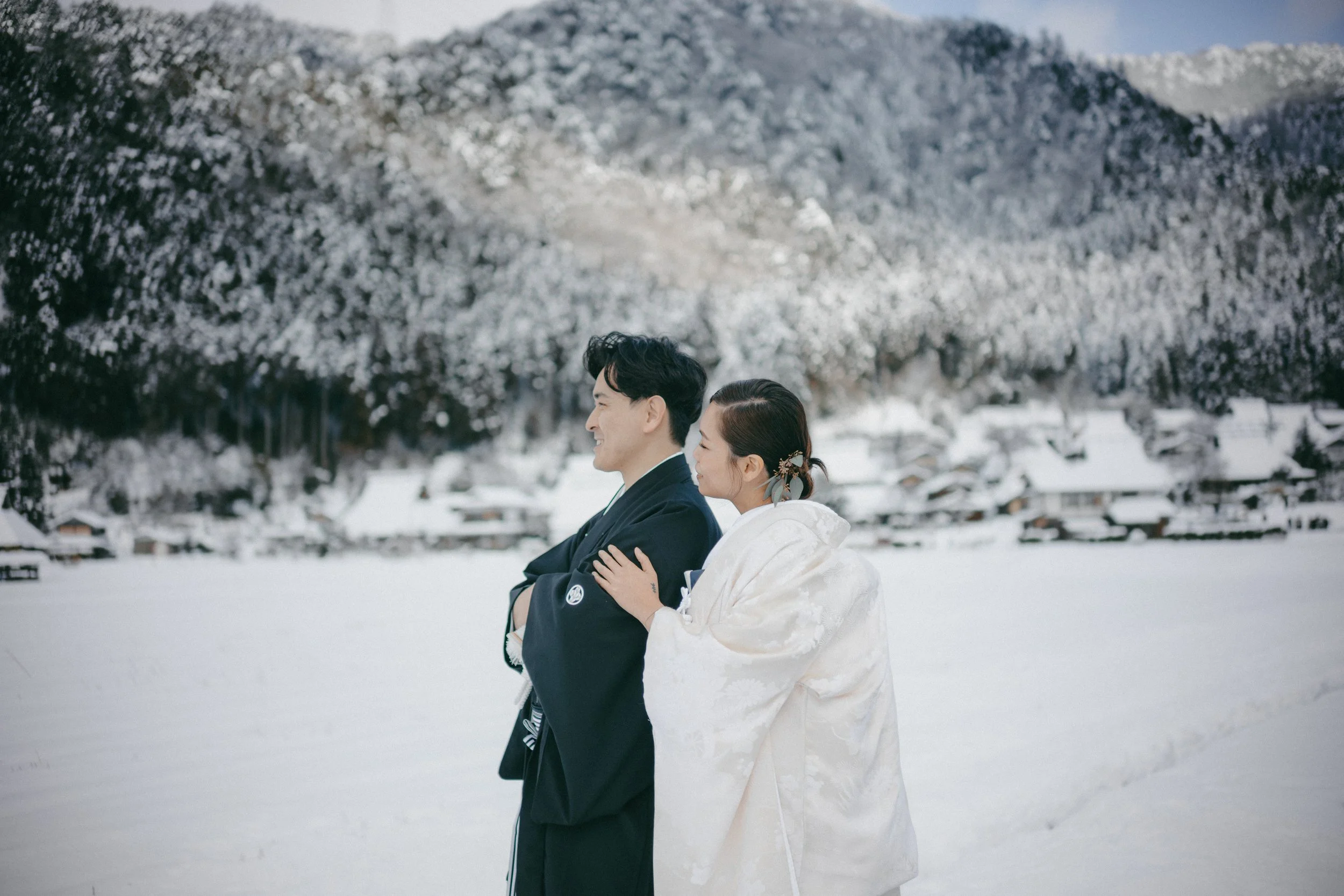 A couple dressed in traditional Japanese wedding attire standing on snow-covered ground with snow-covered mountains and trees in the background.