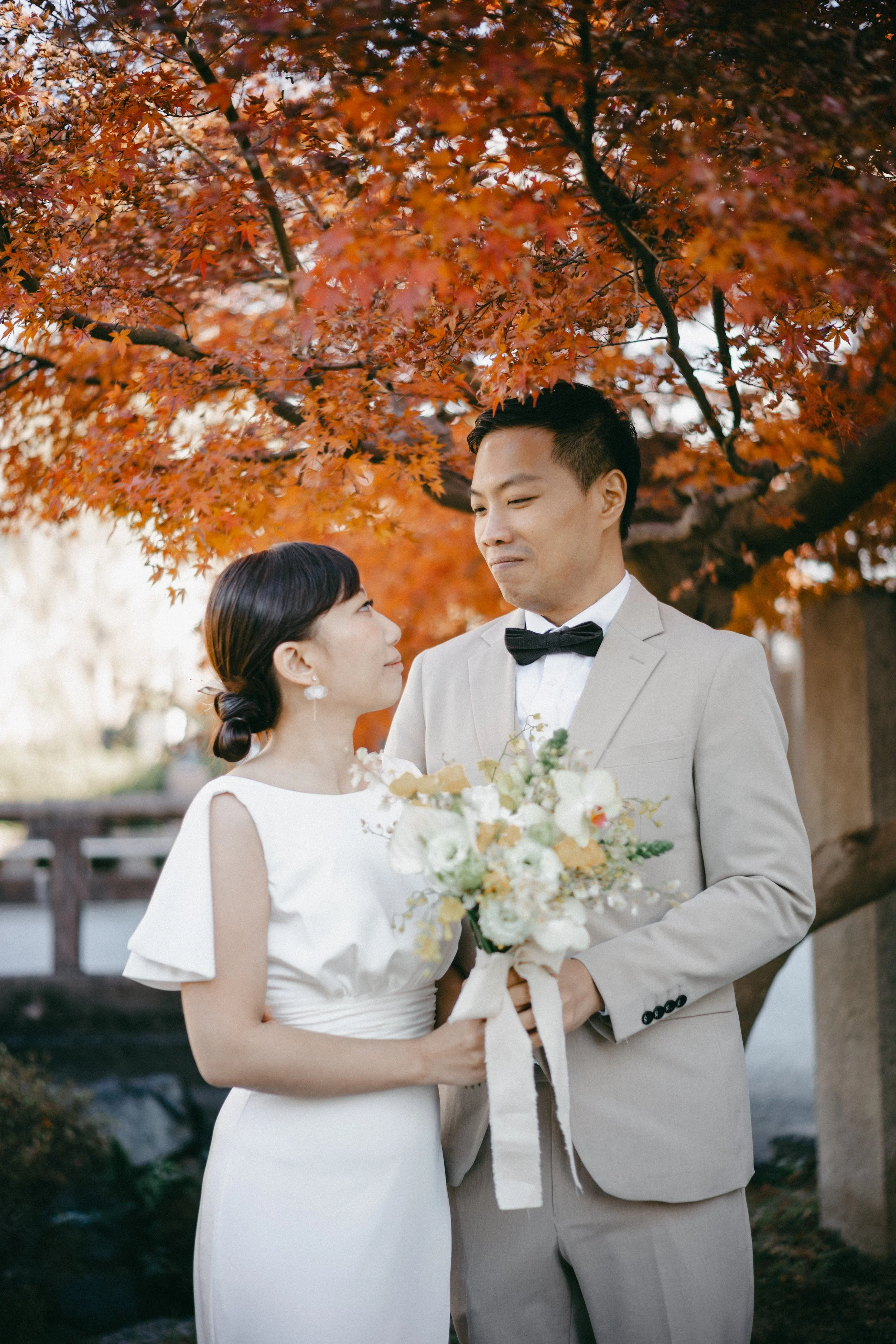 A bride and groom standing outdoors under a tree with vibrant orange leaves, holding a bouquet of flowers, and looking at each other.