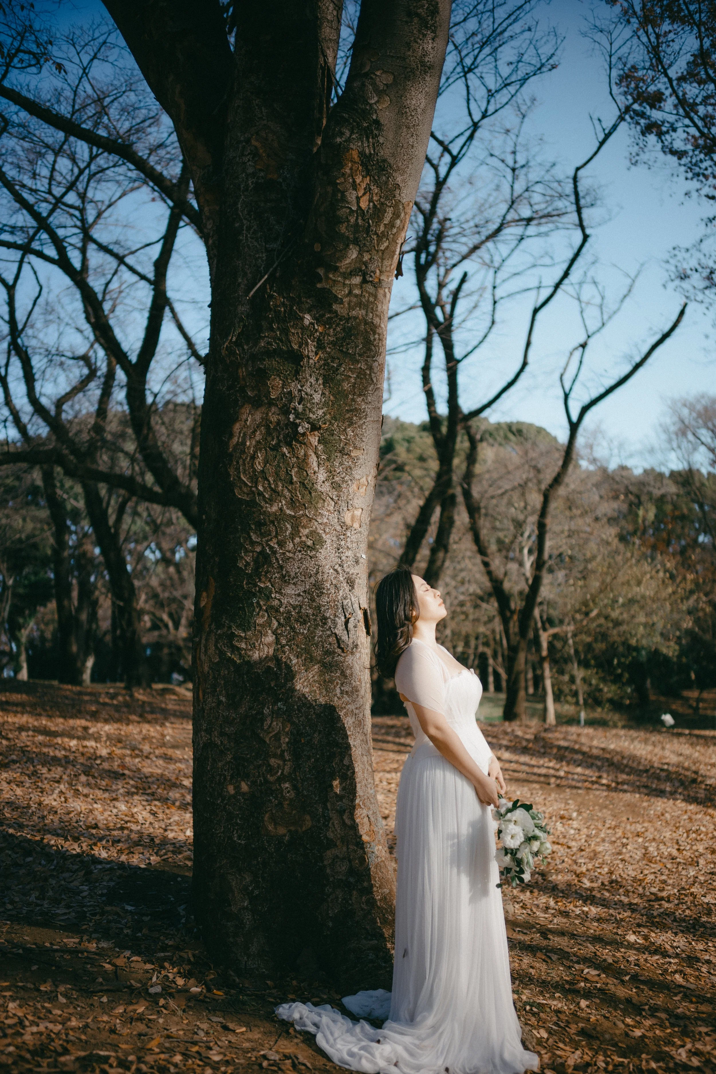 A woman in a white dress holds a bouquet of white flowers and stands outdoors beside a large tree, with her eyes closed and face turned upward in a peaceful pose, during daylight in a wooded area.