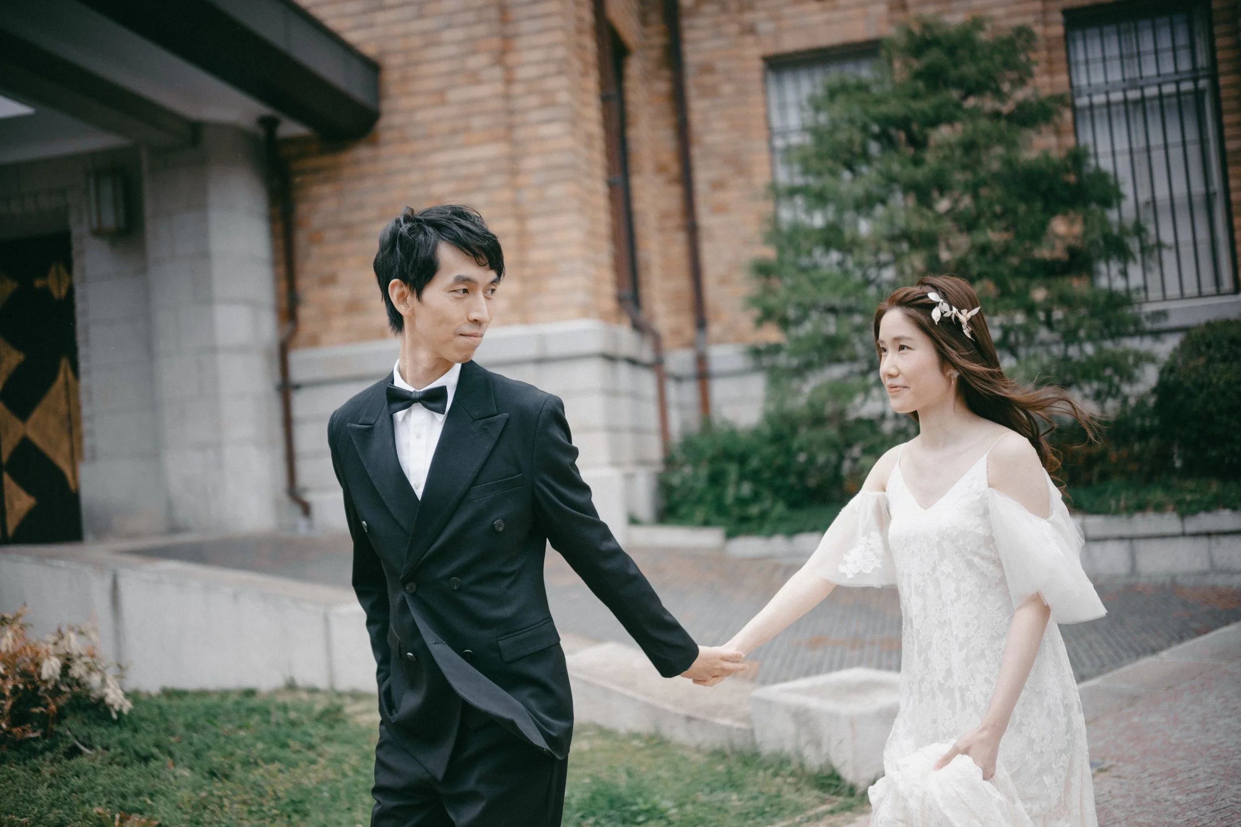 A couple in wedding attire holding hands outdoors, with the man in a black tuxedo and the woman in a lace white wedding dress with cold shoulder sleeves, standing on a city sidewalk near a brick building and greenery.