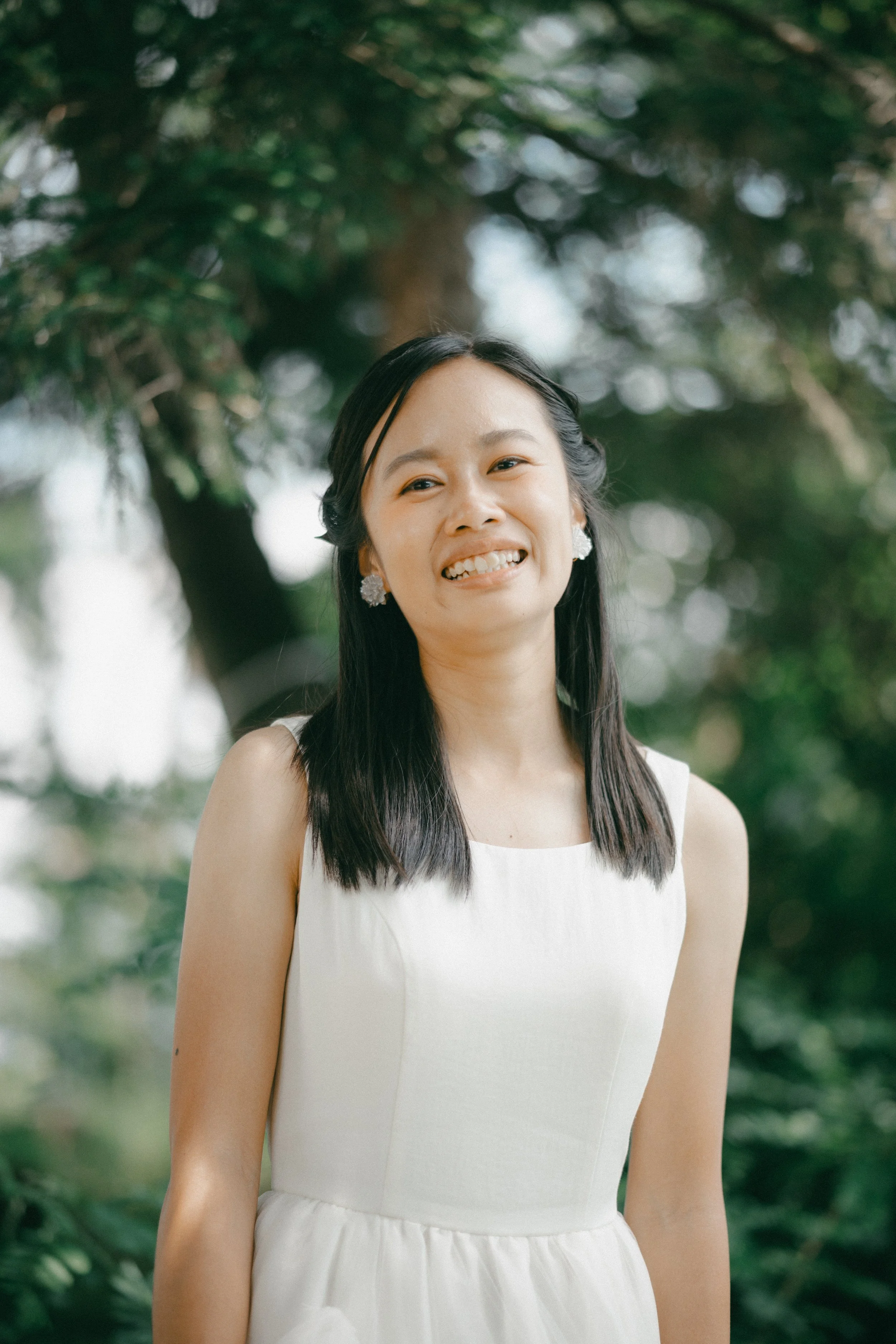 A woman with long black hair, wearing a sleeveless white dress and floral earrings, smiling outdoors with greenery and trees in the background.