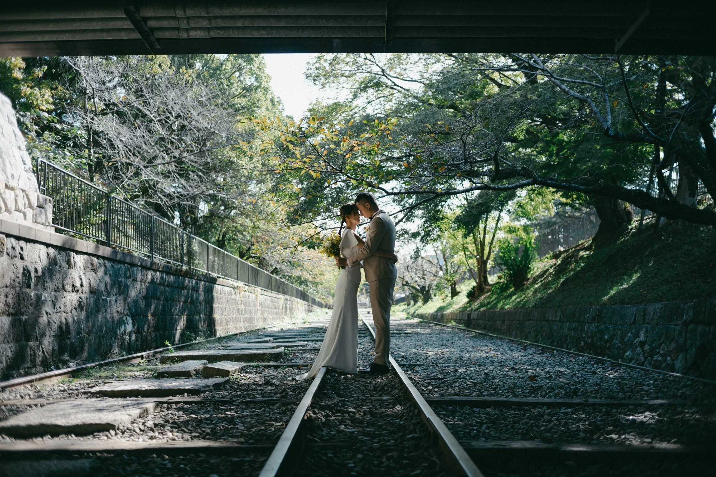 A couple in wedding attire standing on train tracks under a bridge, surrounded by trees with sunlight filtering through.