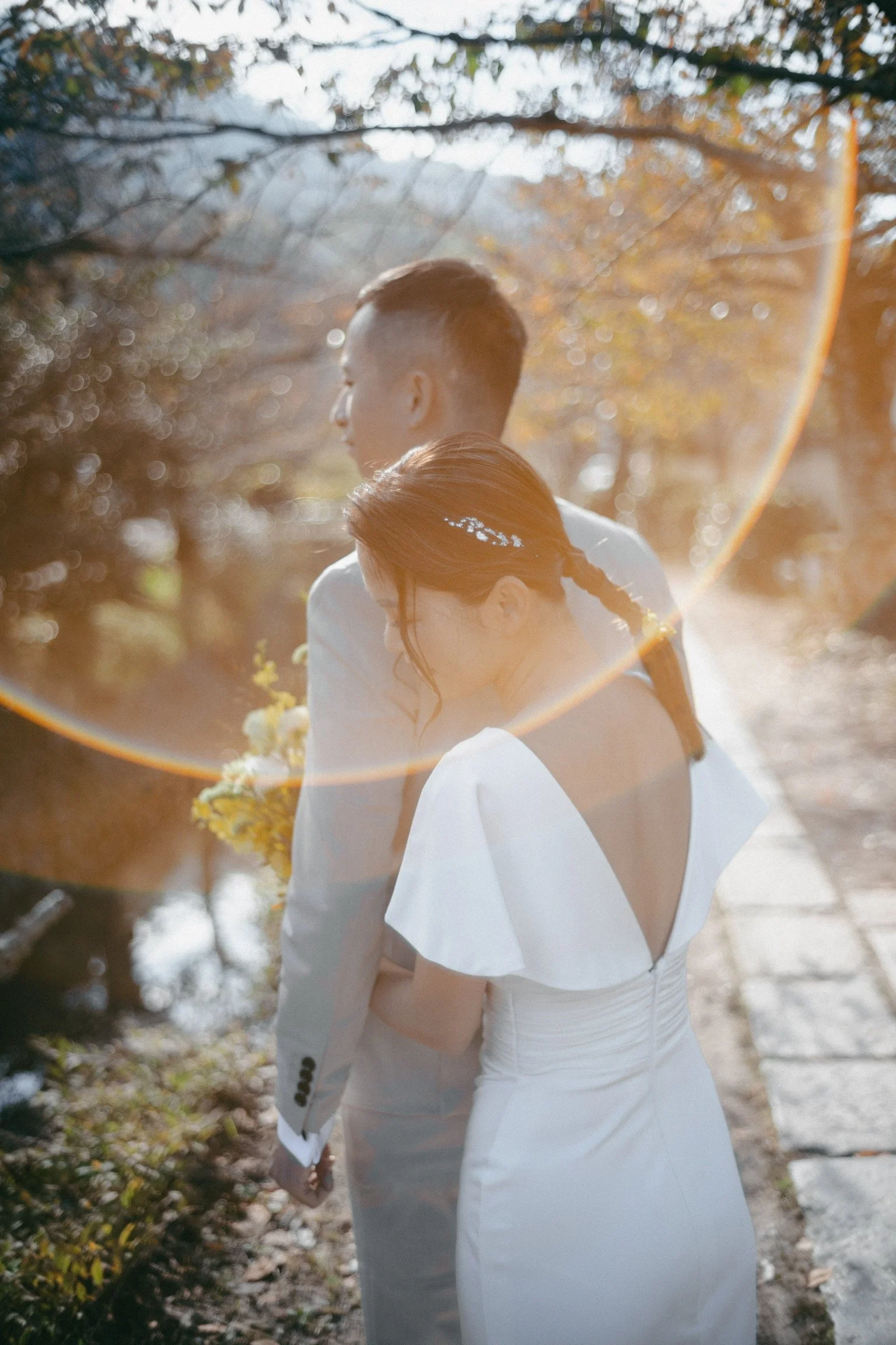 A bride and groom embrace outdoors in a park with autumn foliage, backlit by sunlight creating a lens flare.