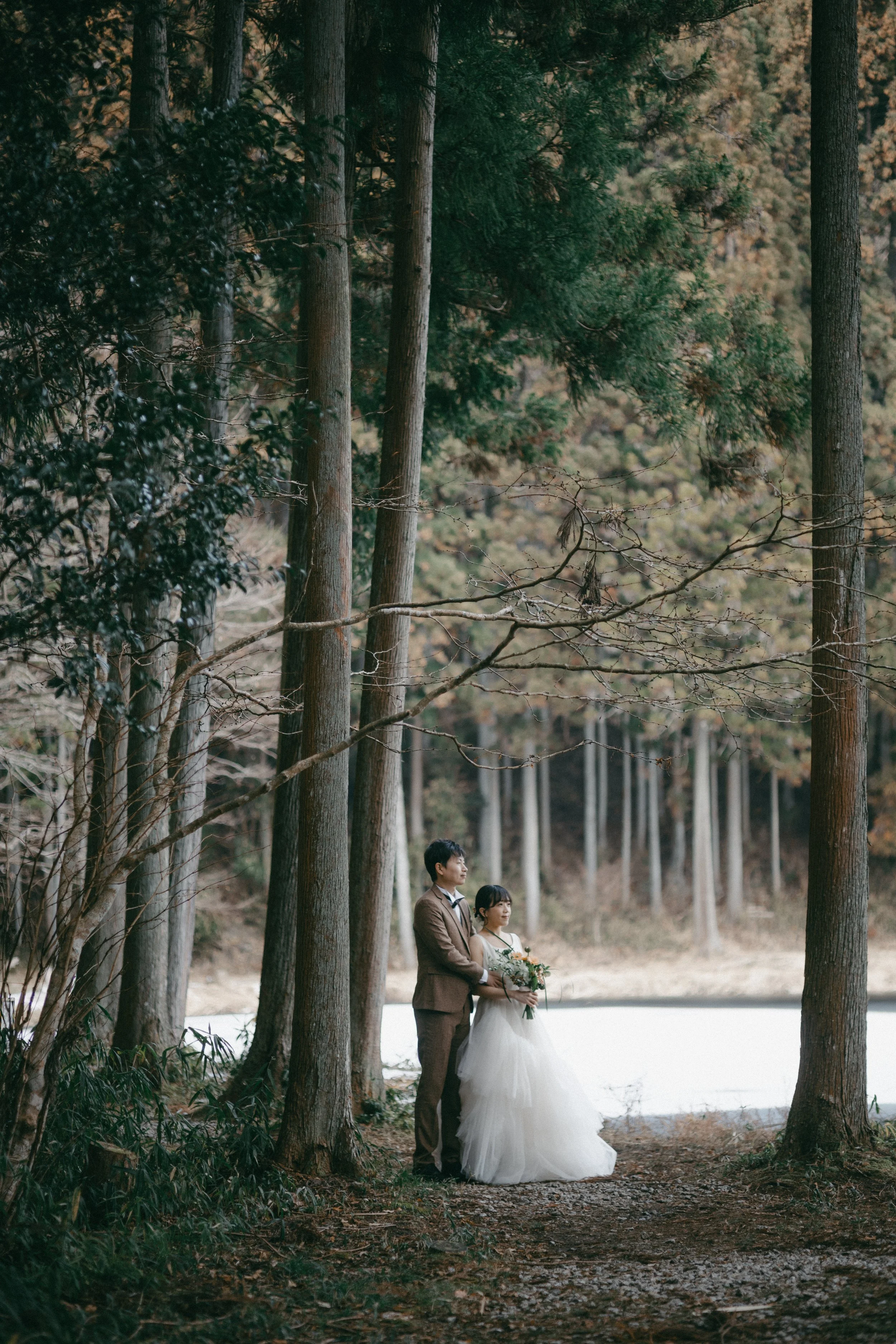 A couple standing in a forest, dressed in wedding attire, with the man in a brown suit and the woman in a white wedding gown, holding a bouquet.
