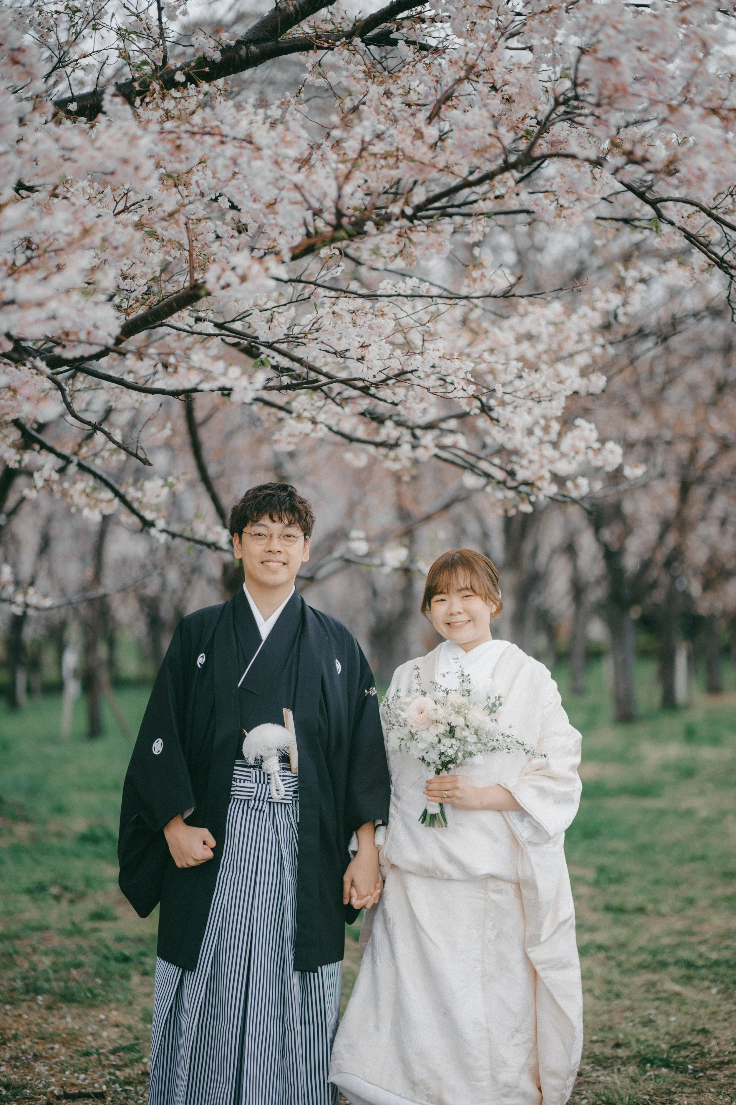 A happy couple in traditional Japanese wedding attire holding hands and smiling under cherry blossom trees during spring.
