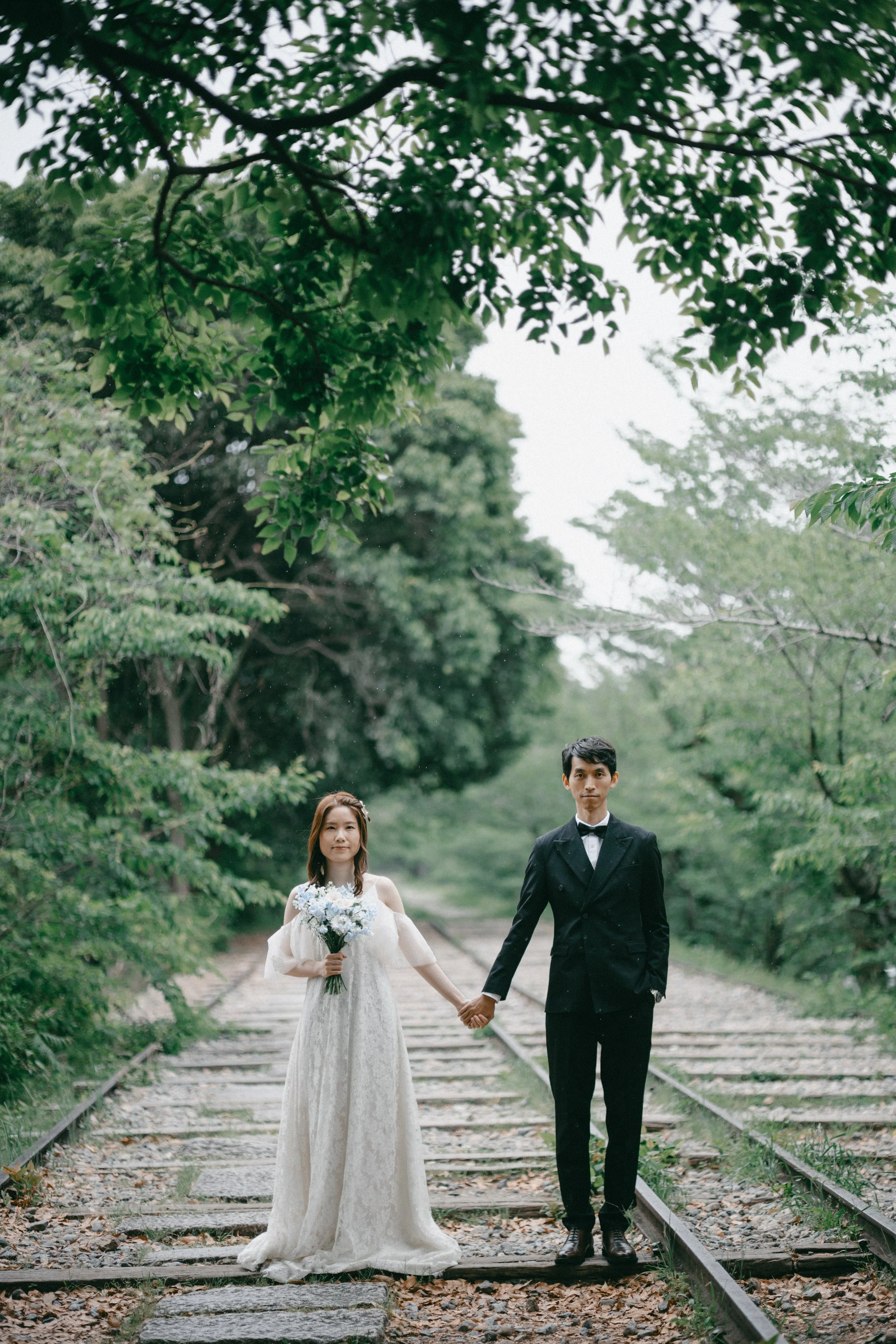 A bride and groom walking hand in hand on a disused train track surrounded by green trees, with the bride holding a bouquet of flowers.