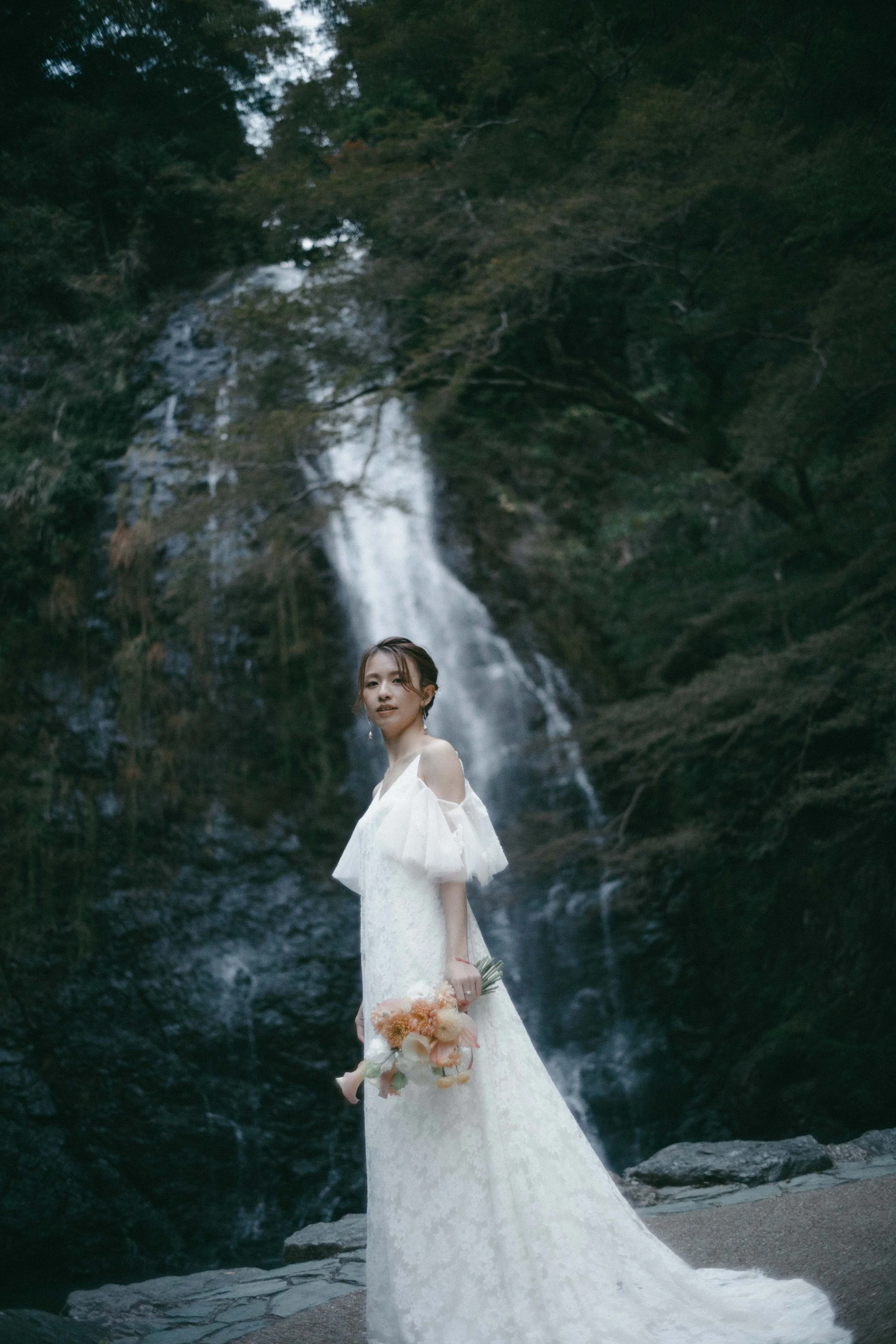A woman in a white wedding dress holding a bouquet in front of a waterfall surrounded by lush greenery.