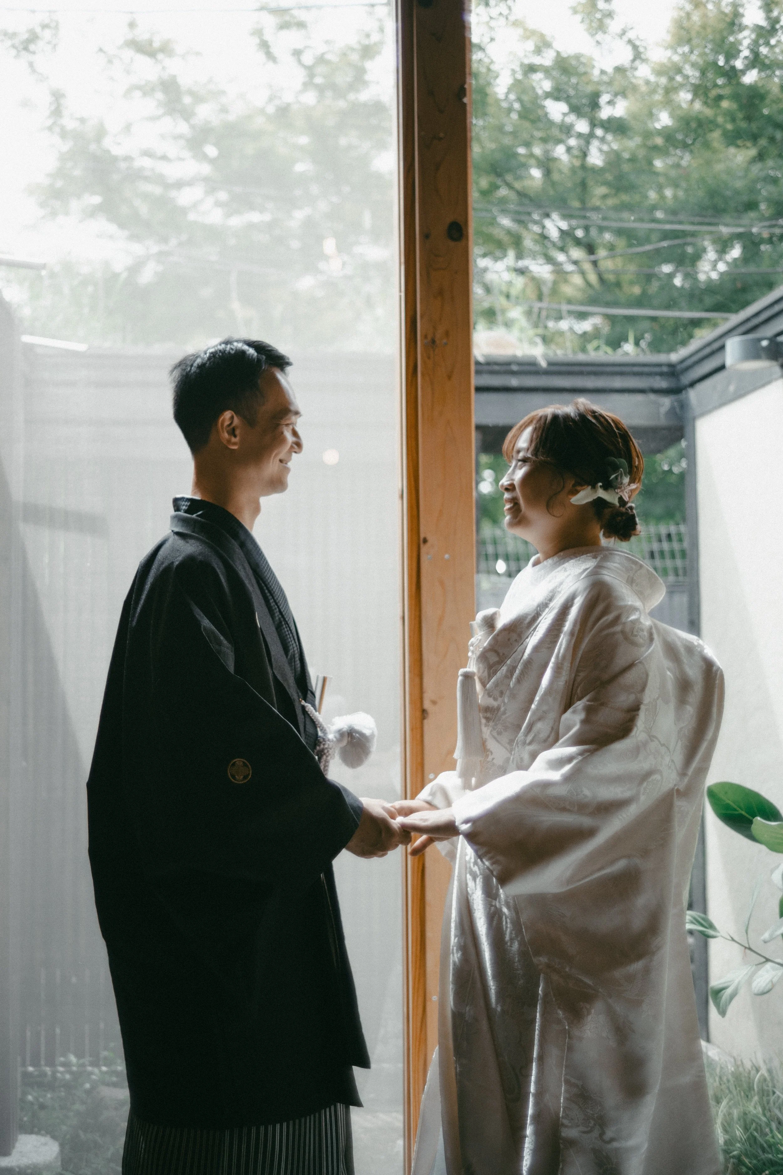 A couple dressed in traditional Japanese wedding attire holding hands and smiling at each other indoors near a large window with greenery outside.