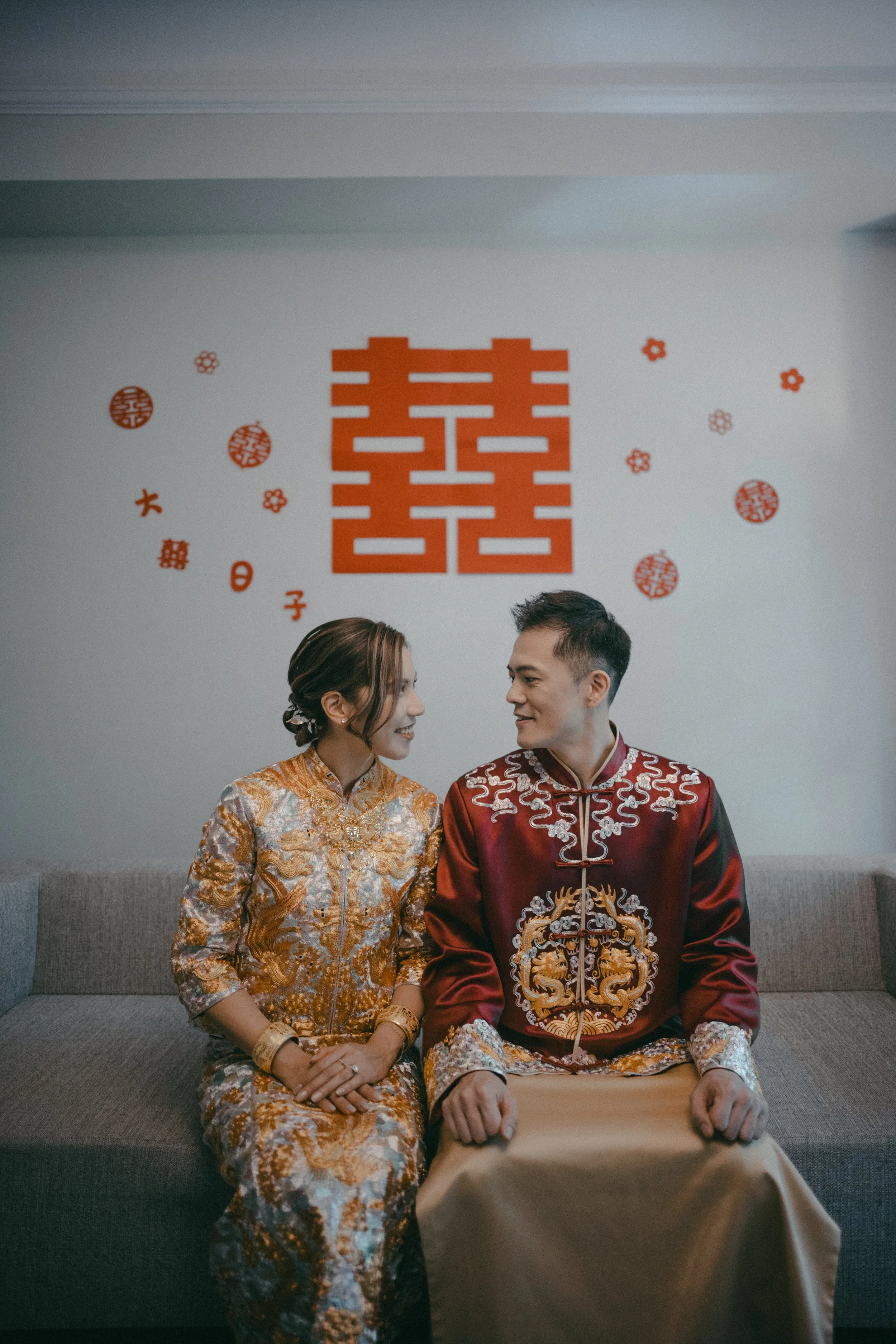 A couple in traditional Chinese wedding attire sitting on a gray couch, smiling at each other, with large red Chinese characters and symbols on the wall behind them.