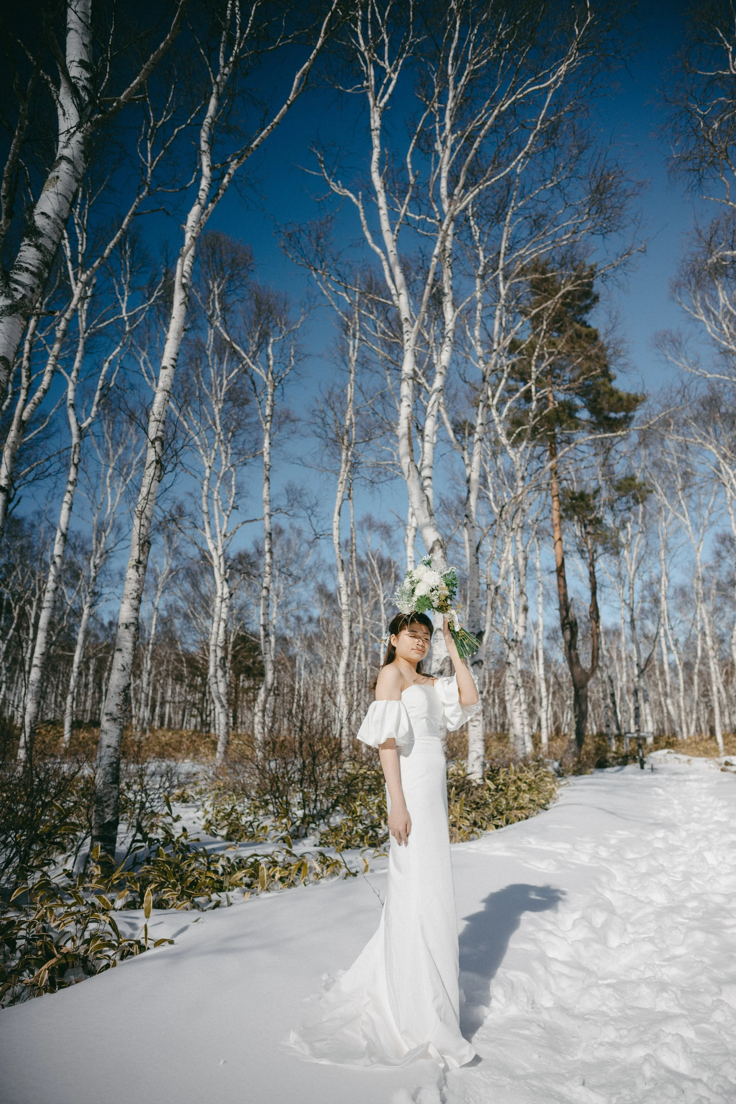 A woman in a white off-shoulder dress holding a bouquet with white flowers, standing in a snowy forest under a clear blue sky.